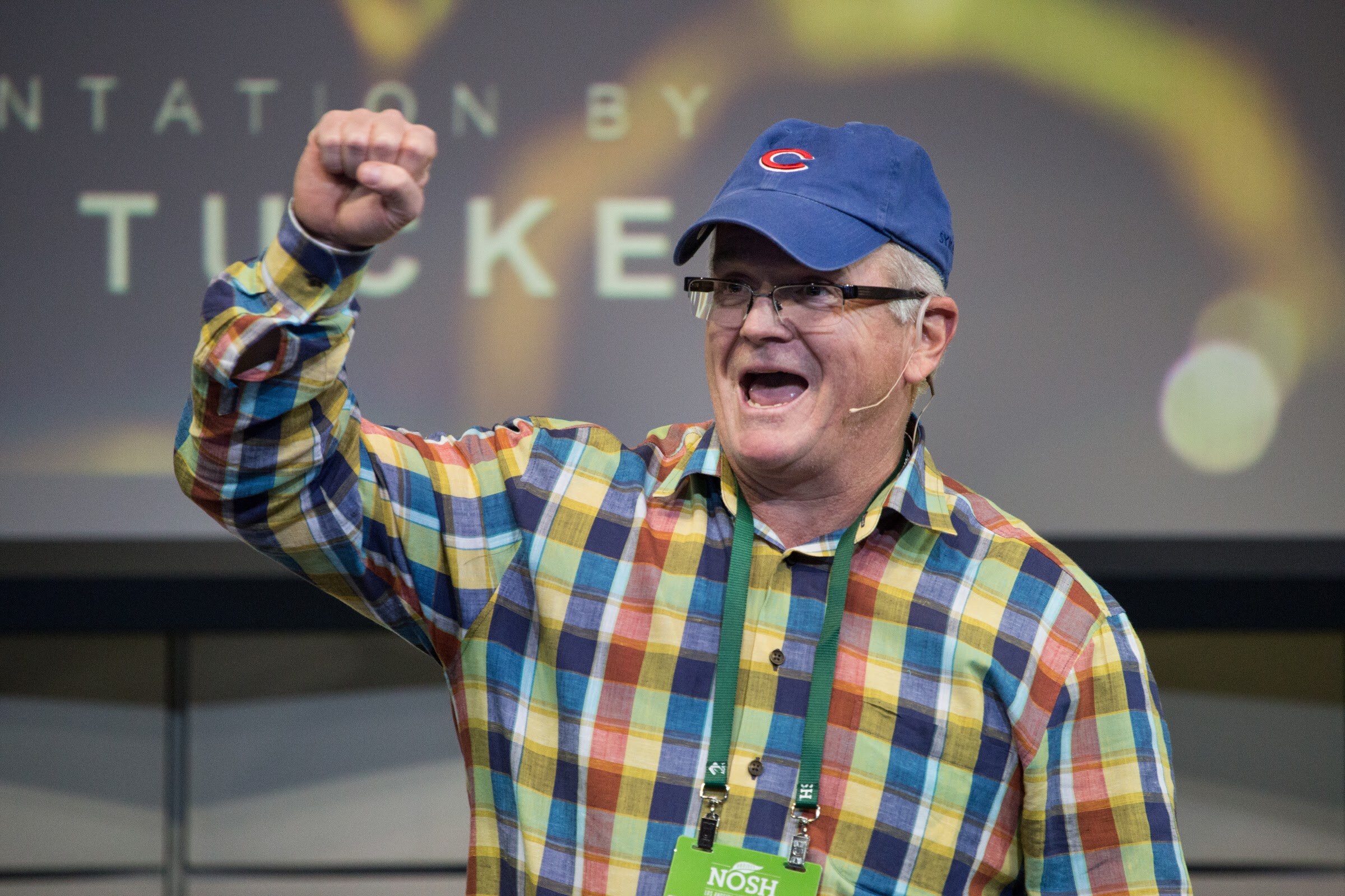 A man in a colorful plaid shirt and blue Cubs cap raises a clenched fist and speaks excitedly in front of a dark projected title slide.