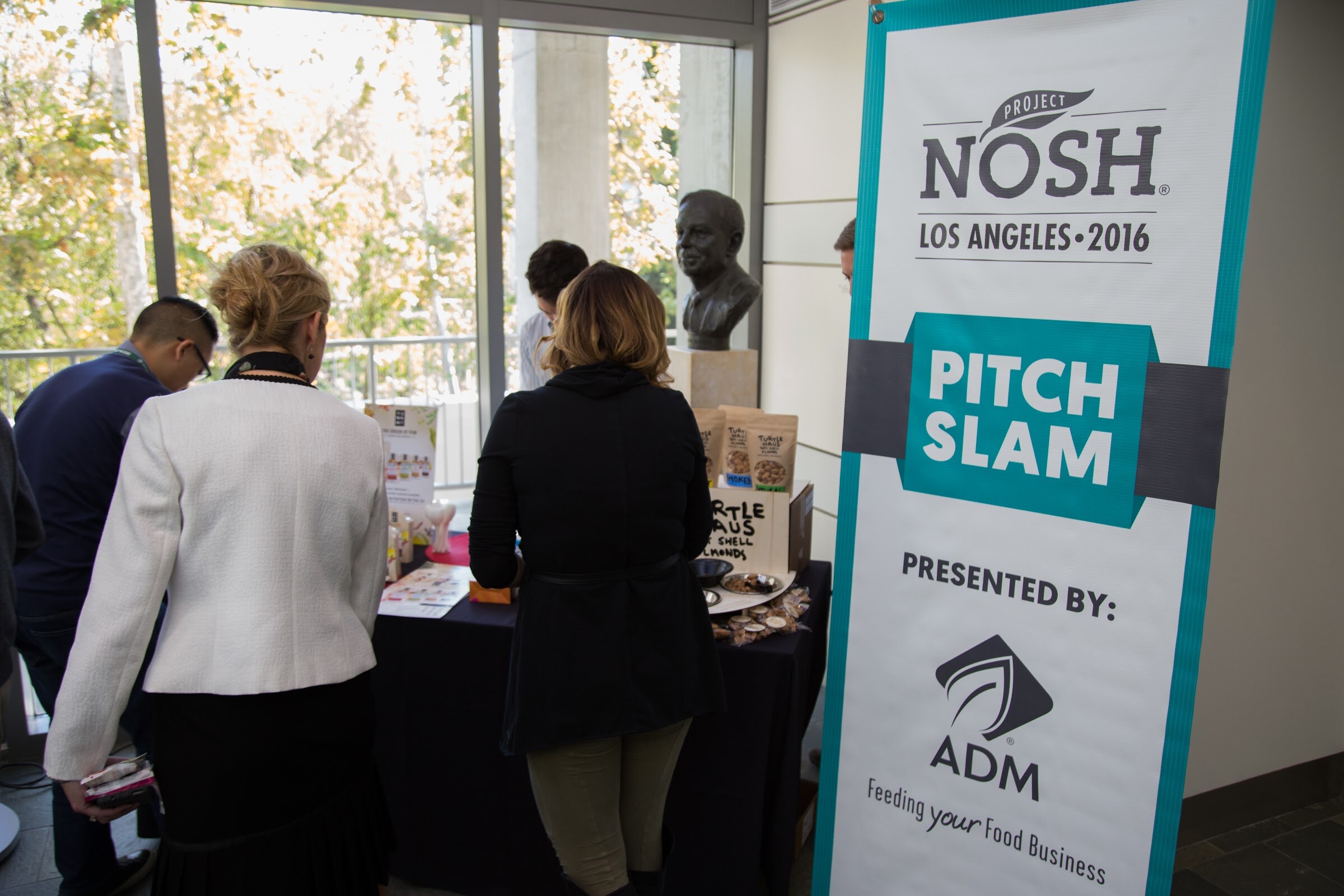 Two women with their backs to the camera look at a table of Turtle Haus almonds and snack samples beside a tall Project NOSH Pitch Slam banner presented by ADM.