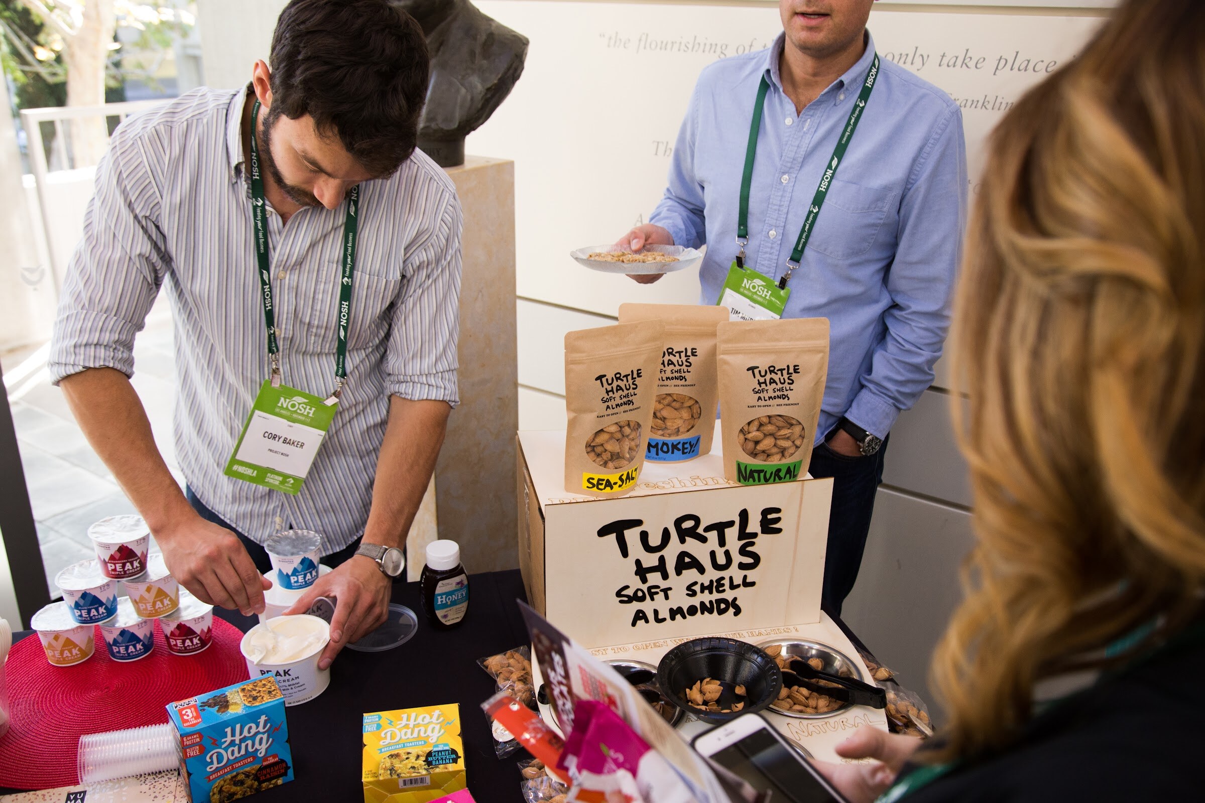 A man in a striped shirt scoops Peak triple cream yogurt while another man in a blue shirt holds a plate behind a display of Turtle Haus soft shell almonds; both wear NOSH badges.