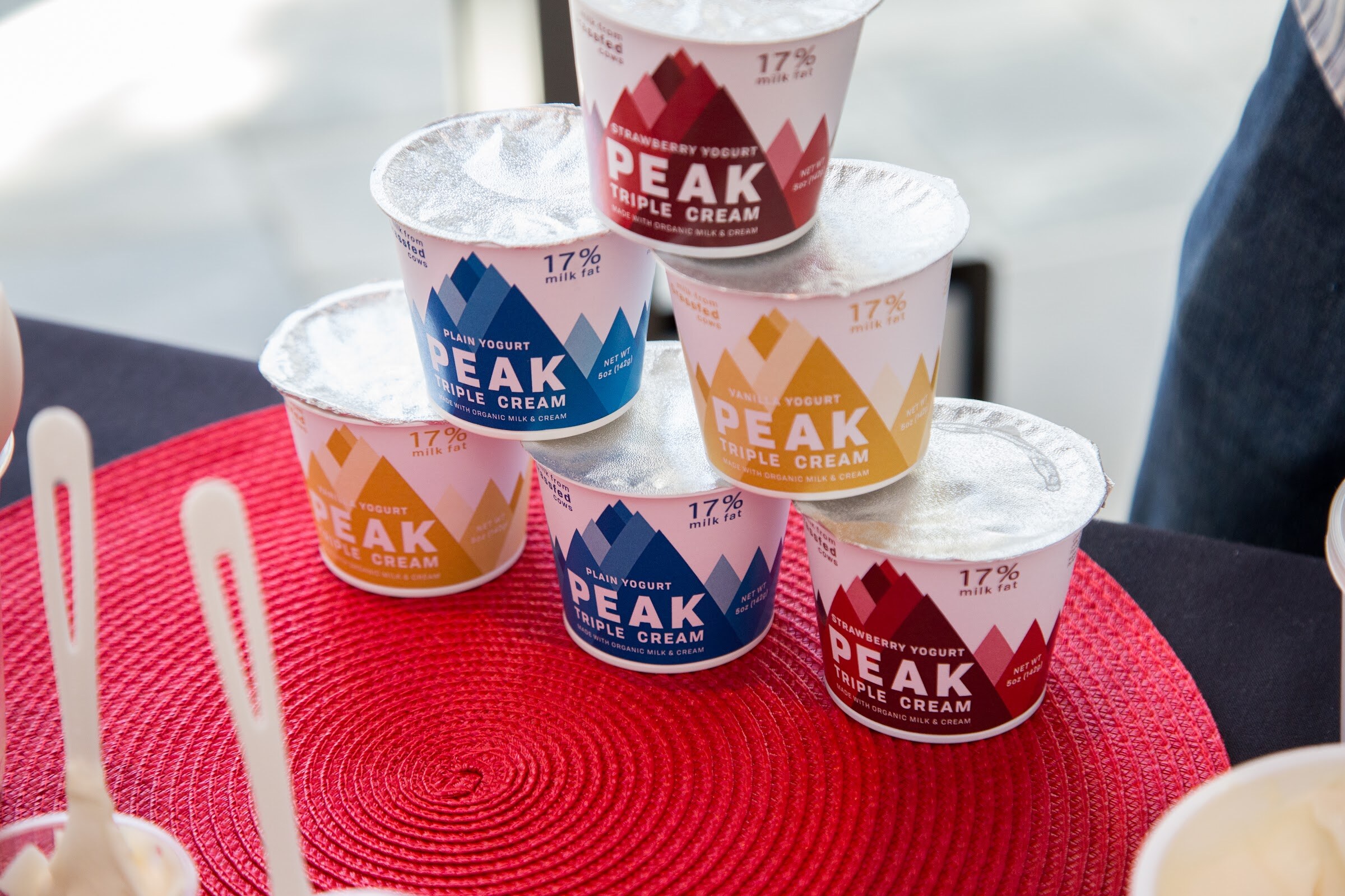 A pyramid of Peak Triple Cream yogurt cups with strawberry, plain and vanilla labels sits on a red woven placemat next to plastic sample spoons.