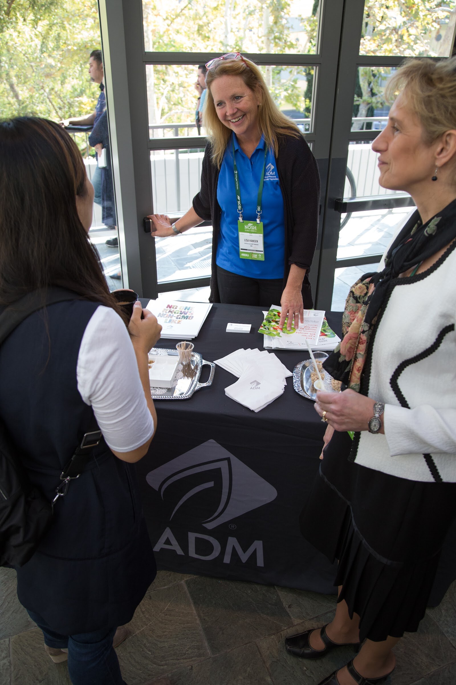 A woman in a blue ADM polo and NOSH badge smiles behind a black table cloth printed with the ADM logo as two guests face her, with printed brochures and a tray of samples on the table.