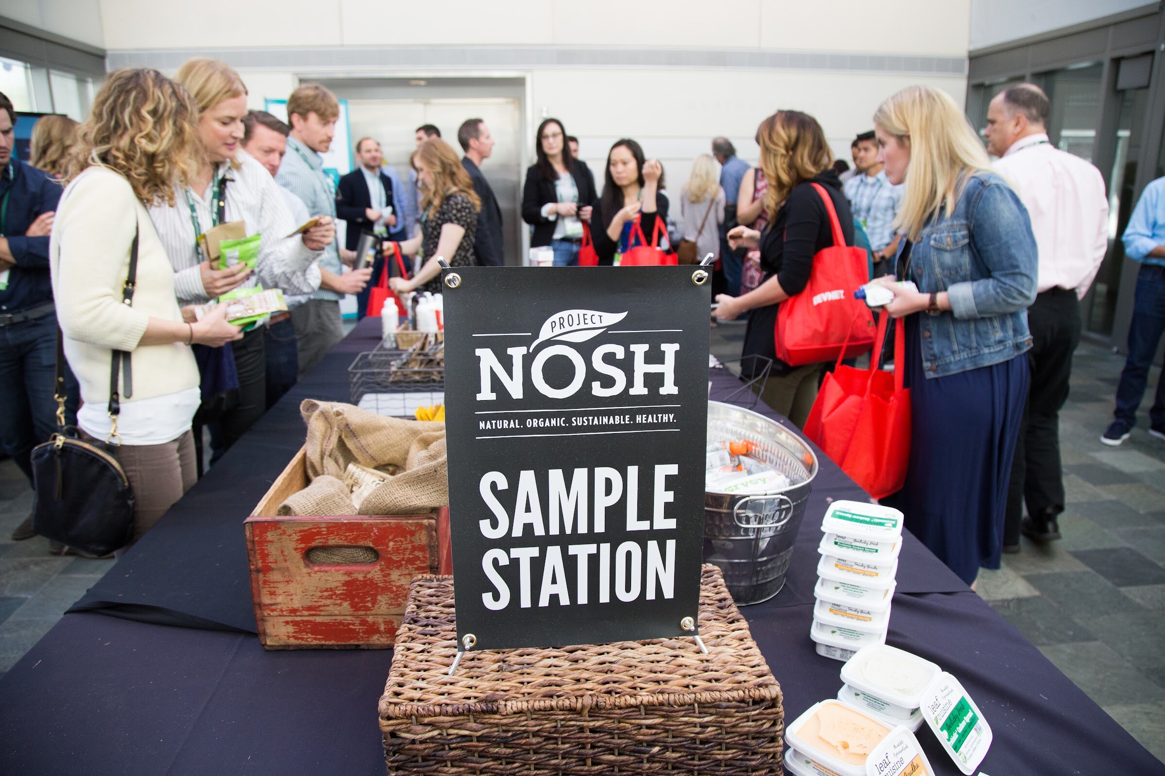 A black Project NOSH Sample Station sign sits on a basket on a long table with leaf cuisine spread tubs, while attendees carrying red tote bags stand and chat behind it.