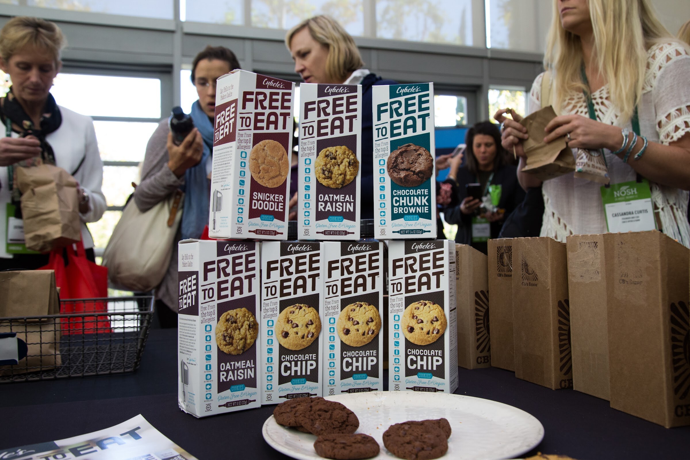 Two rows of Cybele's Free to Eat cookie boxes in oatmeal raisin, chocolate chip, snickerdoodle and chocolate chunk brownie flavors, with a plate of cookies in front and attendees holding paper bags behind.