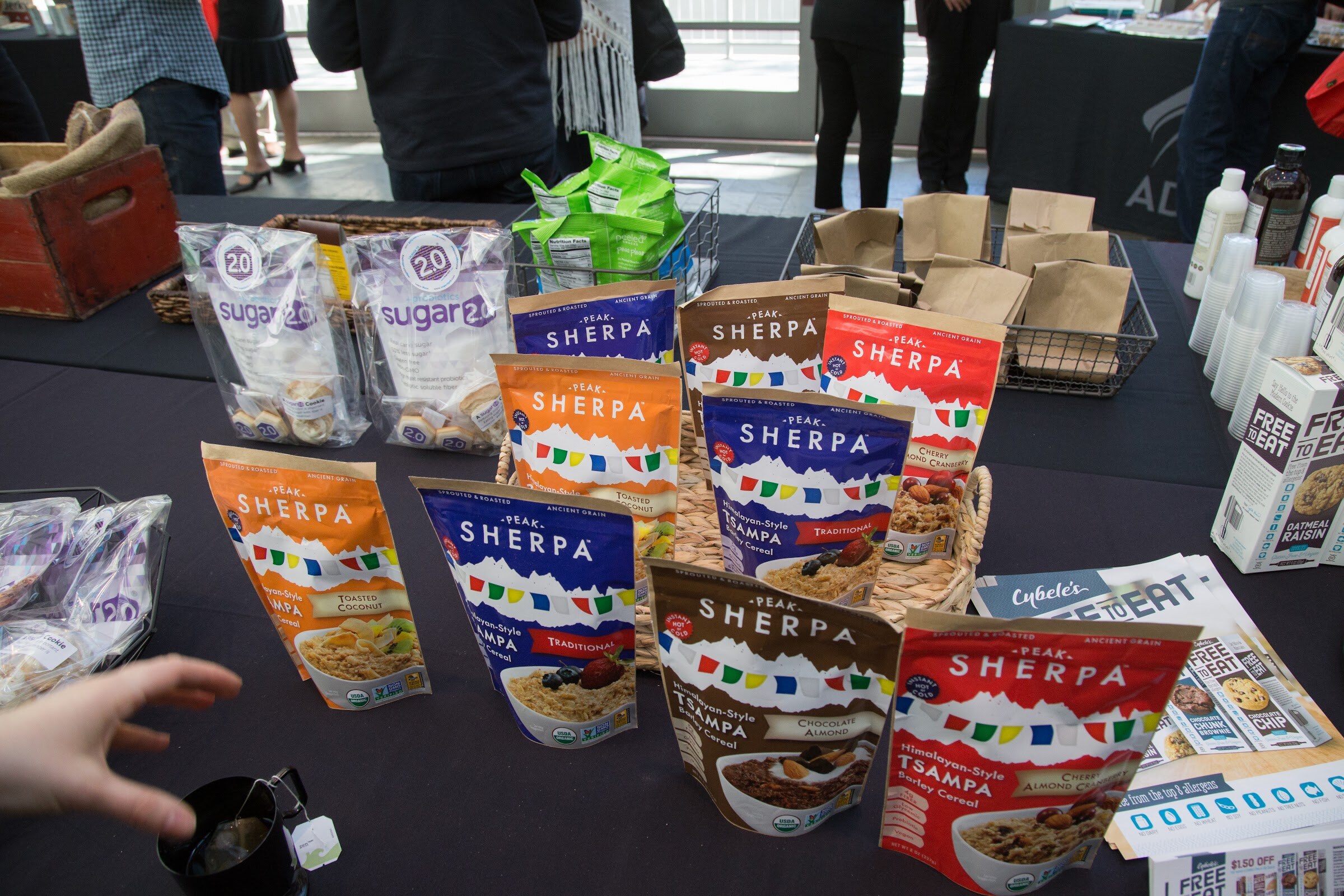Orange, blue, brown and red Peak Sherpa tsampa barley cereal pouches fan out in front of a basket on a black table, with Sugar 2.0 bags at left and Cybele's Free to Eat coupons at right.