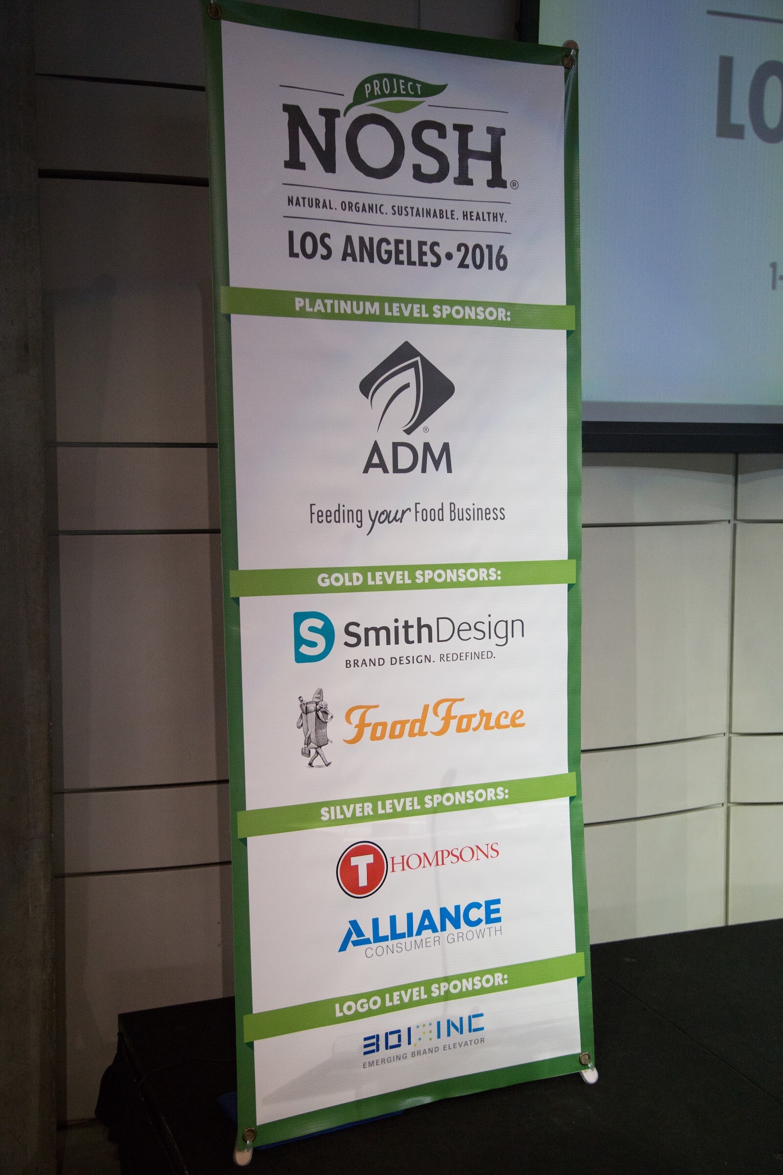 A tall green-edged banner reading Project NOSH Los Angeles 2016 with Platinum sponsor ADM, Gold sponsors SmithDesign and Food Force, Silver sponsors Thompsons and Alliance Consumer Growth, and a logo-level sponsor, standing on a stage.