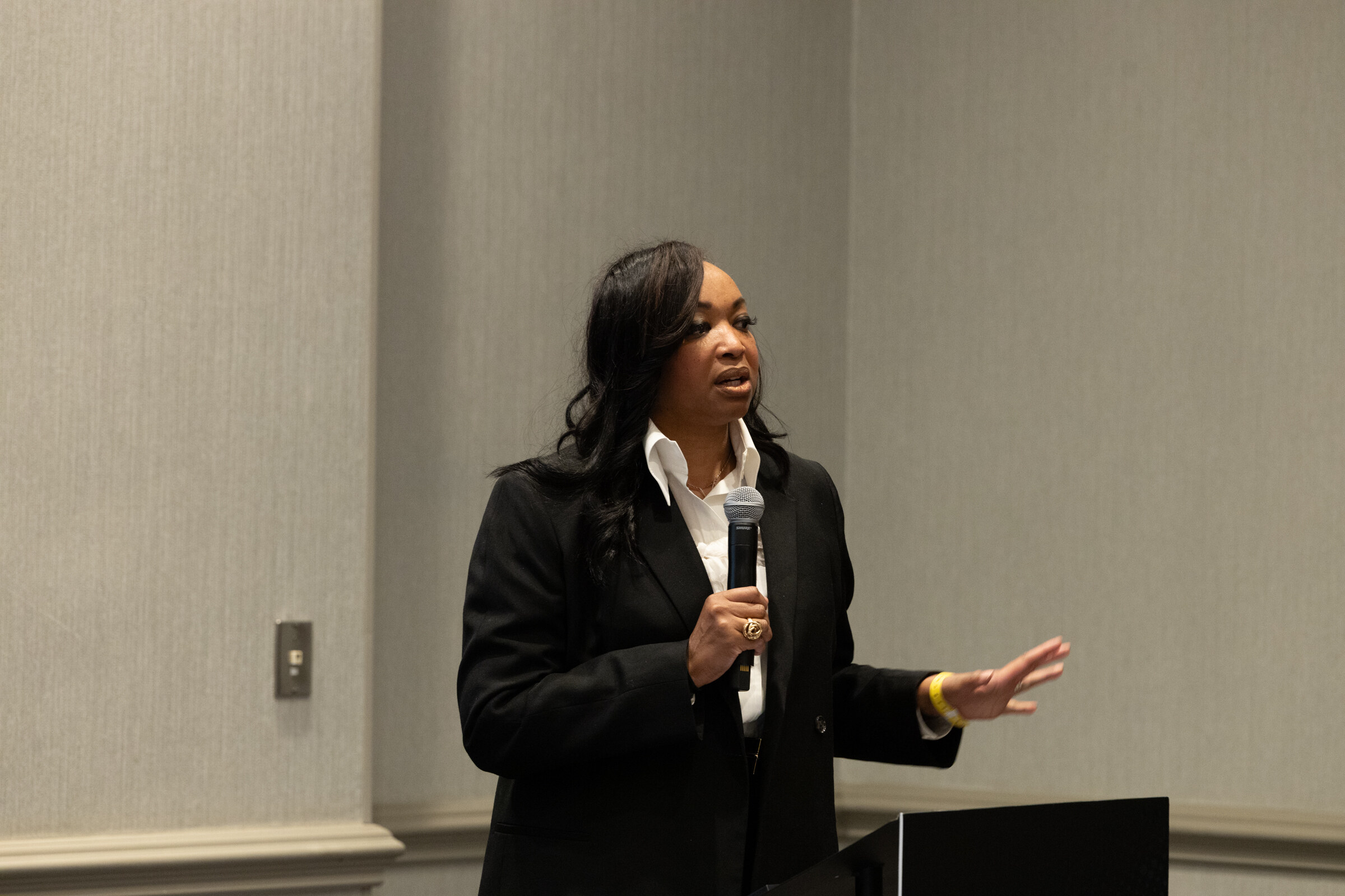 A woman in a black jacket holding a microphone and speaking against a plain wall.