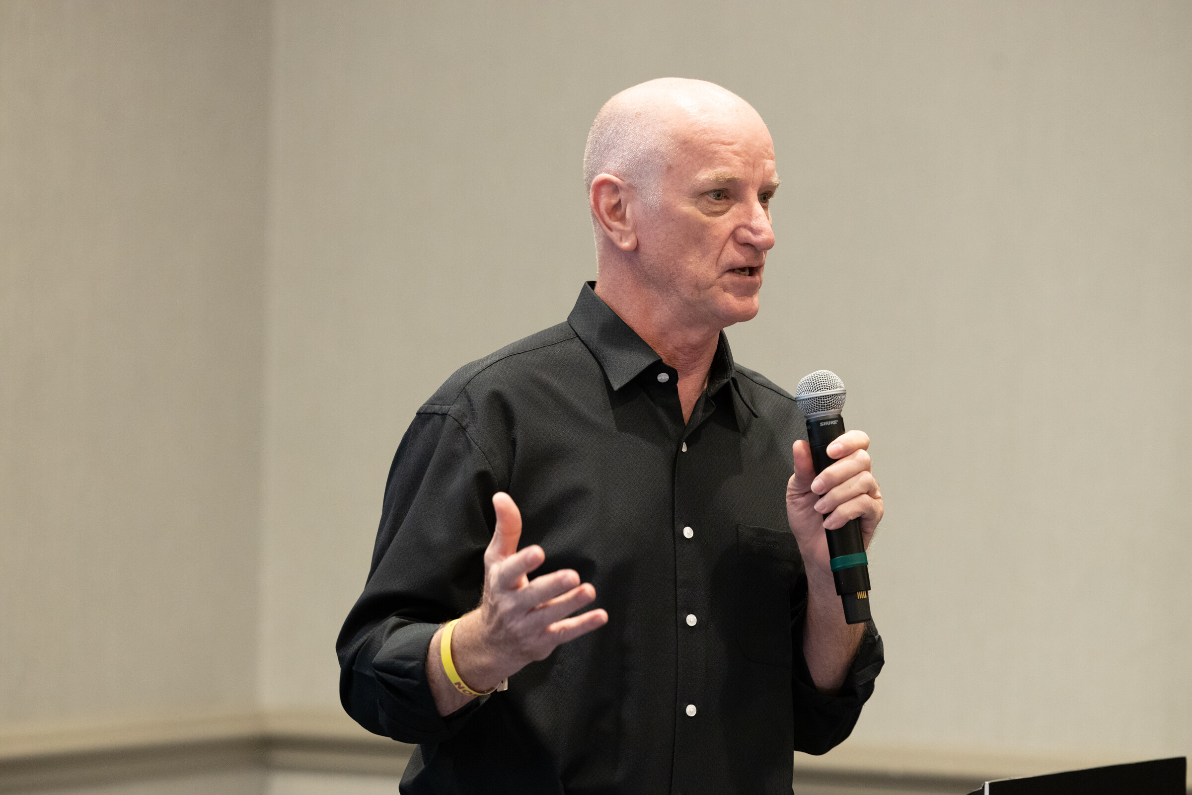 A bald man in a black shirt holding a microphone and gesturing with one hand.