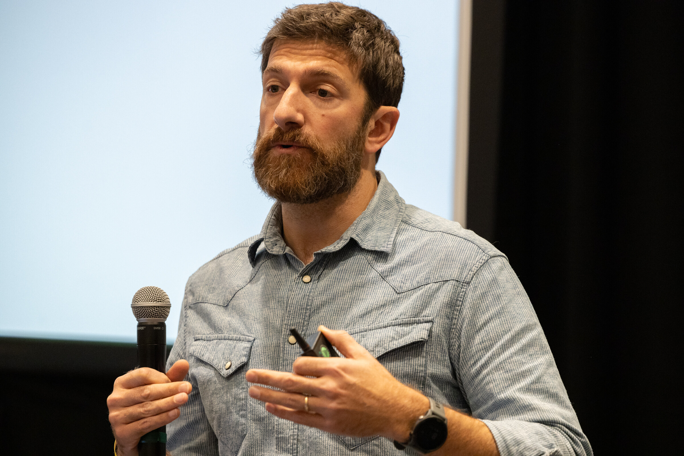A bearded man in a gray denim shirt holding a microphone and a clicker.