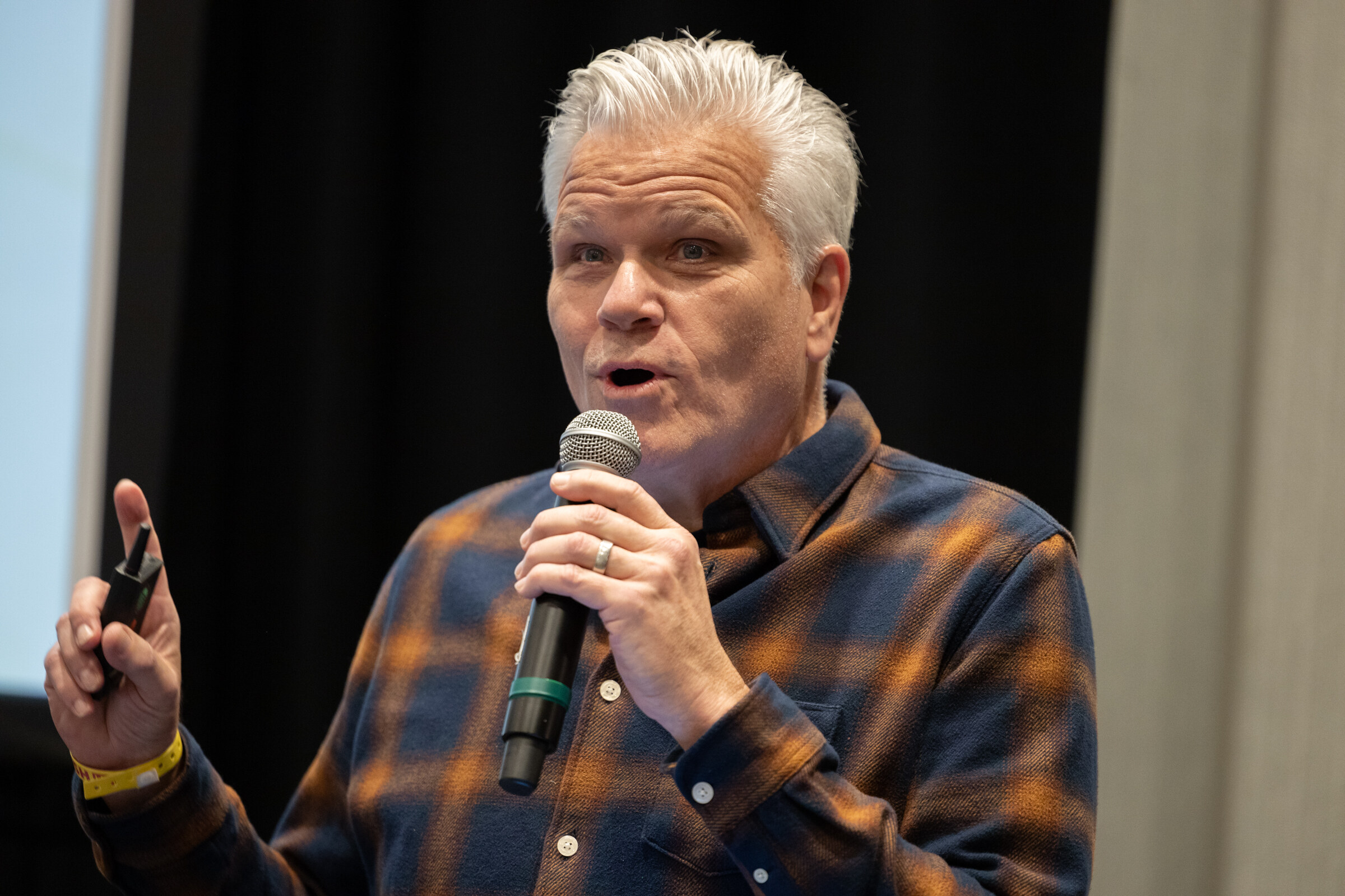 A gray-haired man in a plaid flannel shirt holding a microphone and pointing upward as he speaks.
