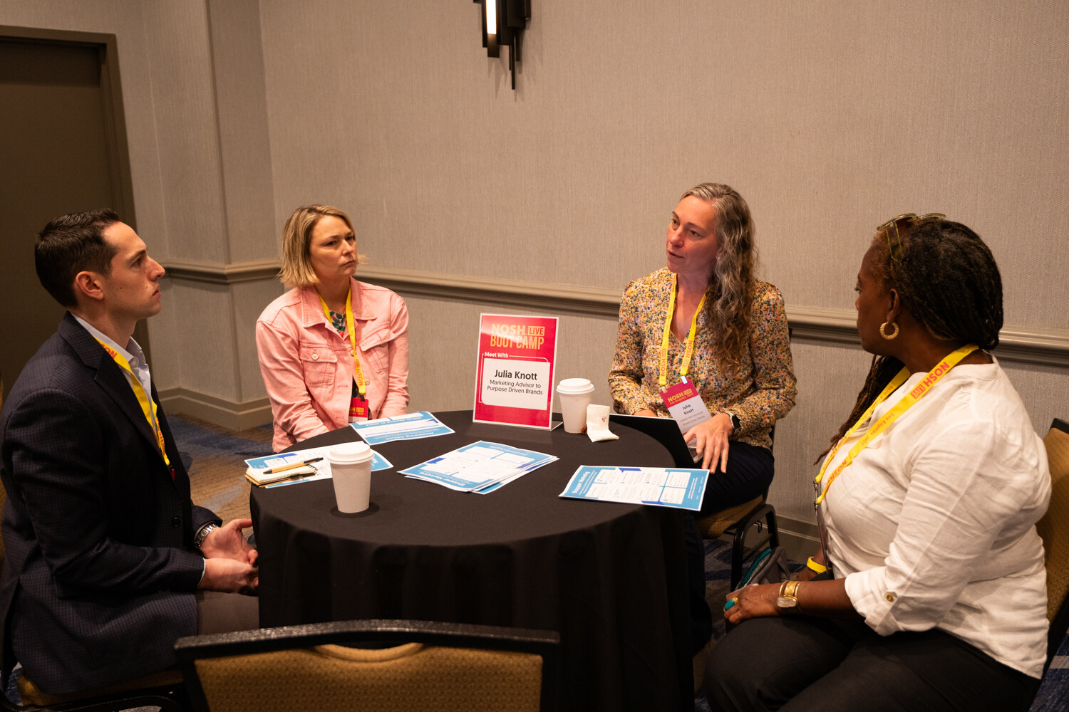 Four people seated around a round table with worksheets and coffee cups, a table sign standing in the middle.