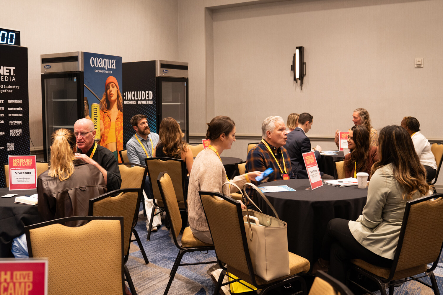 A meeting room with several round tables of people in conversation and a Coaqua banner and refrigerators at the back.