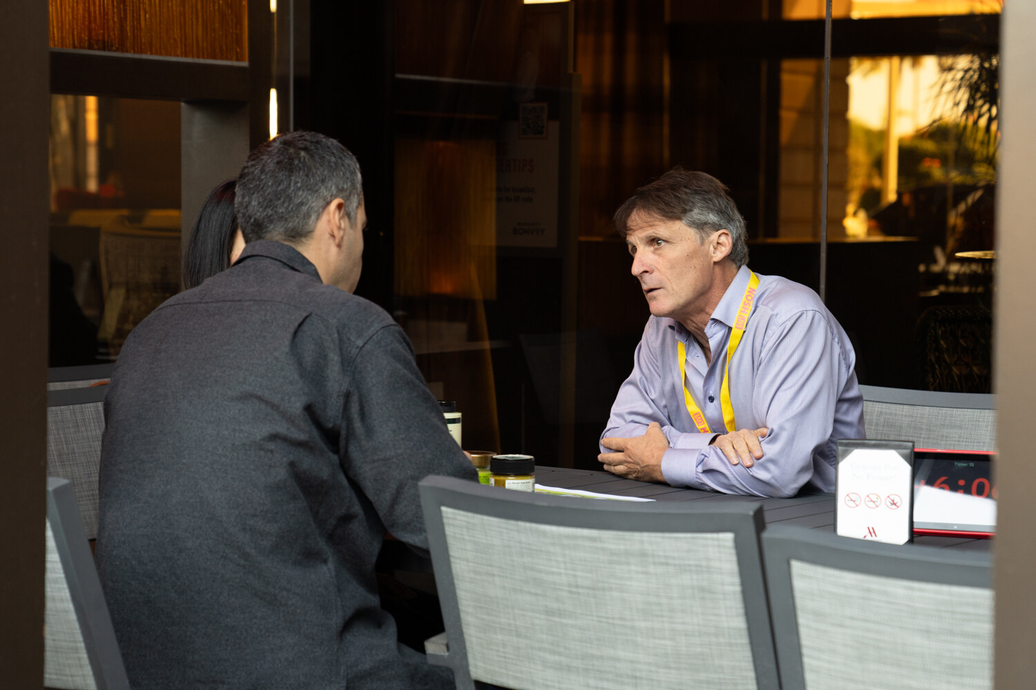 A man in a lavender shirt and lanyard speaking across a small table from a man seen from behind.