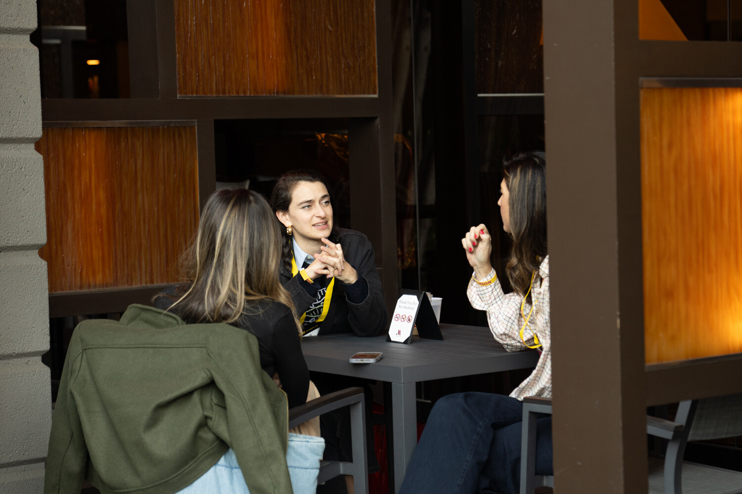Three people seated at a small table in the outdoor lounge, one with hands clasped and a phone on the table.
