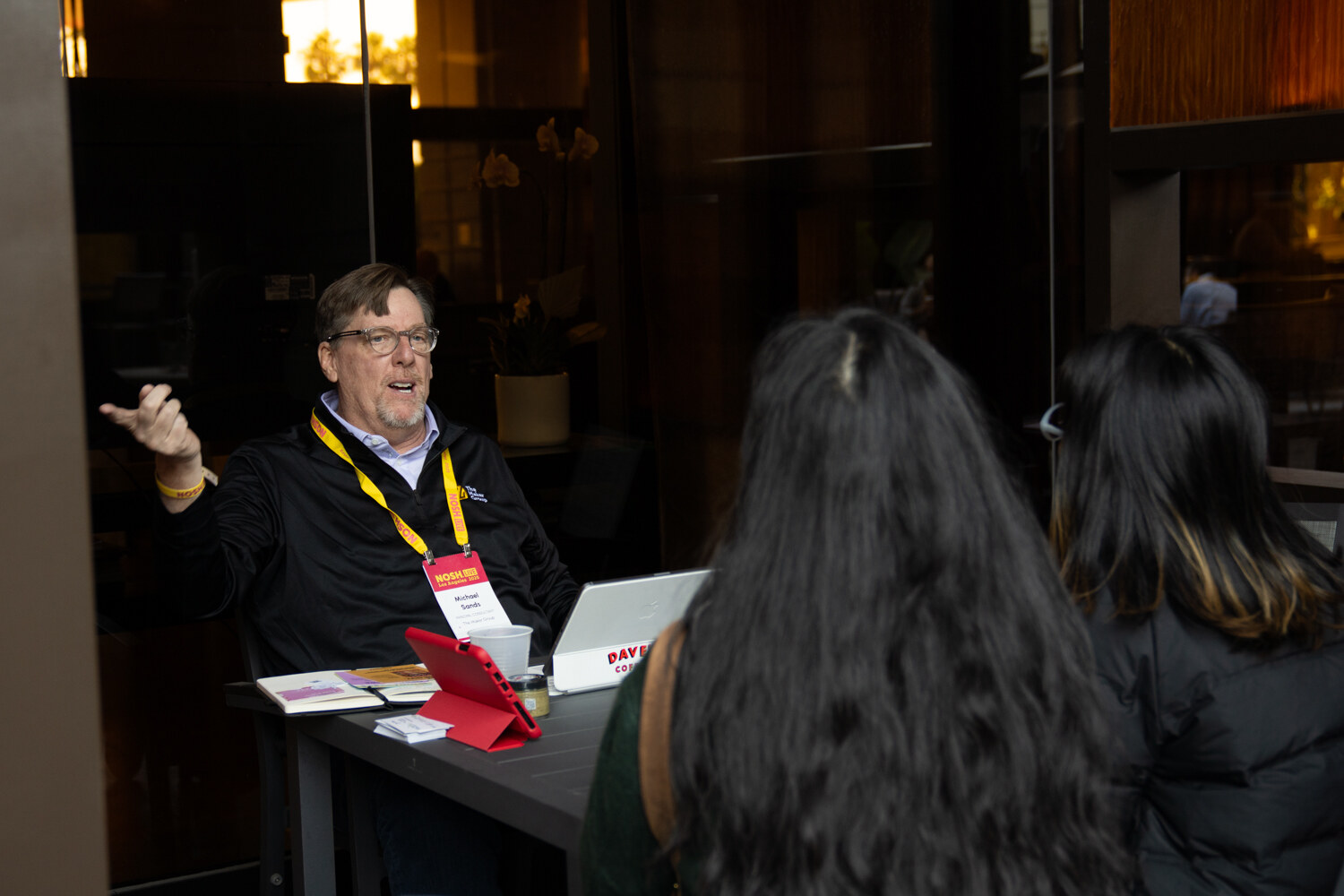 A man in a black jacket and glasses at a table with a laptop in front of two people seen from behind.