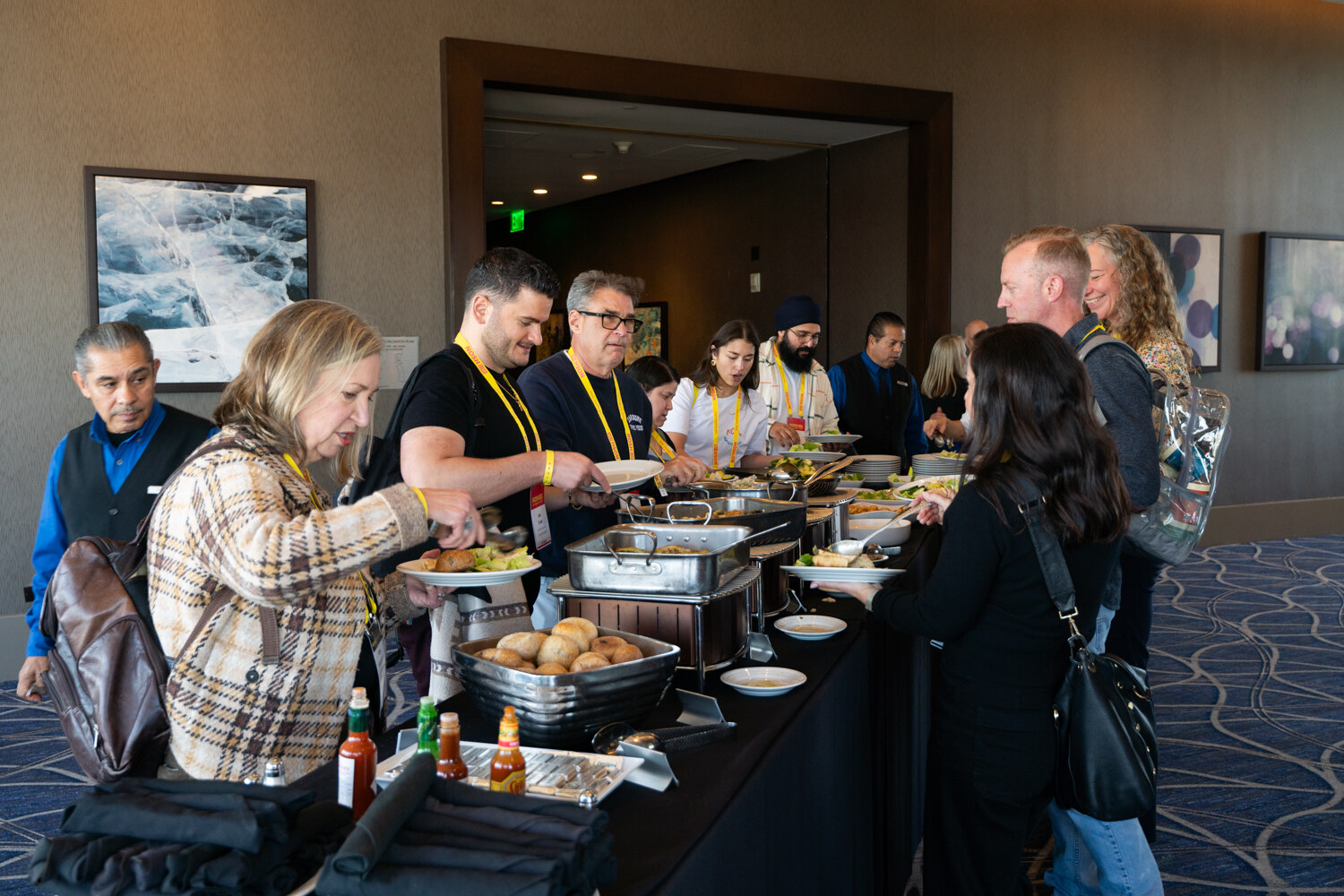 A line of attendees with plates at a long buffet table with chafing dishes.
