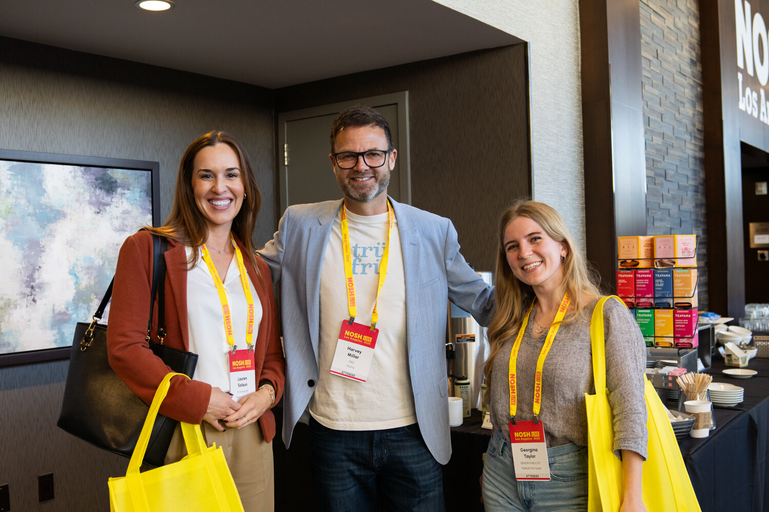 Two women and a man in lanyards smiling in front of a hotel coffee station with yellow tote bags.