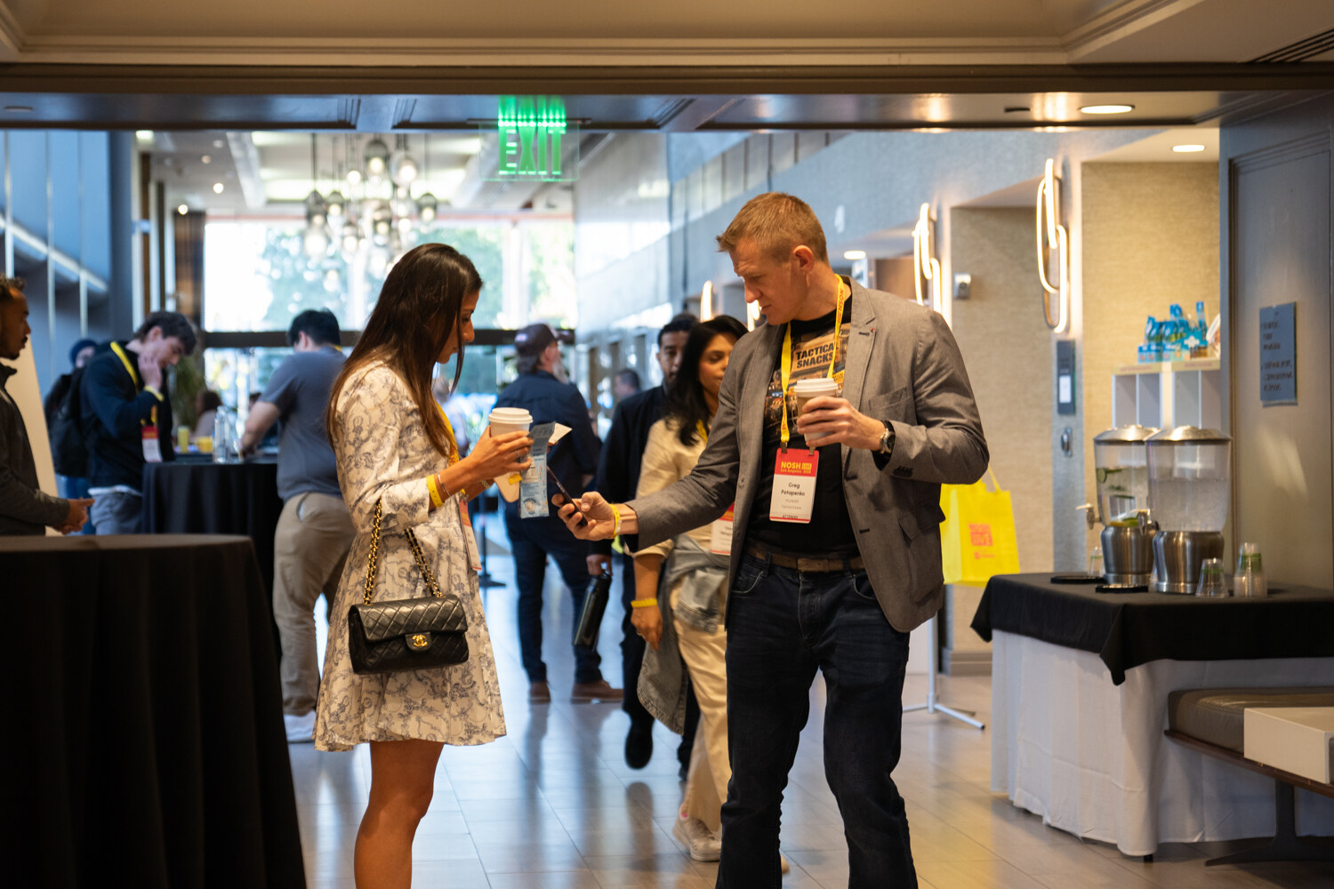A woman and a man in a gray blazer holding coffee in a bright hotel corridor.