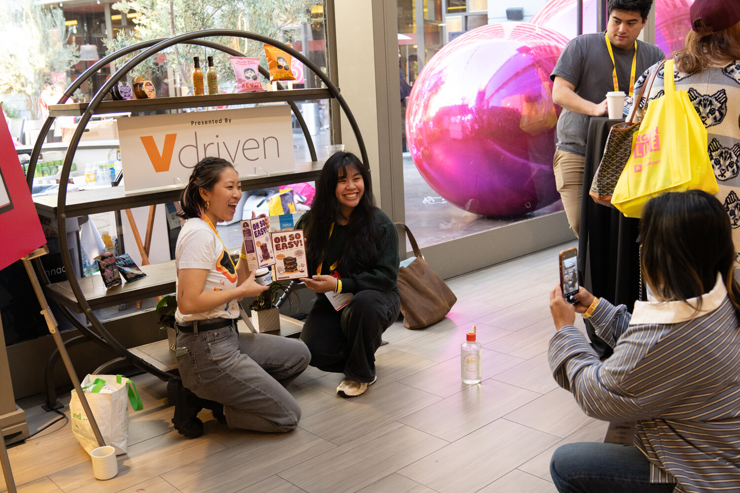 Two women kneeling by a round shelf with a Vdriven sign, one holding boxes of Oh So Easy, a third person taking a photo.
