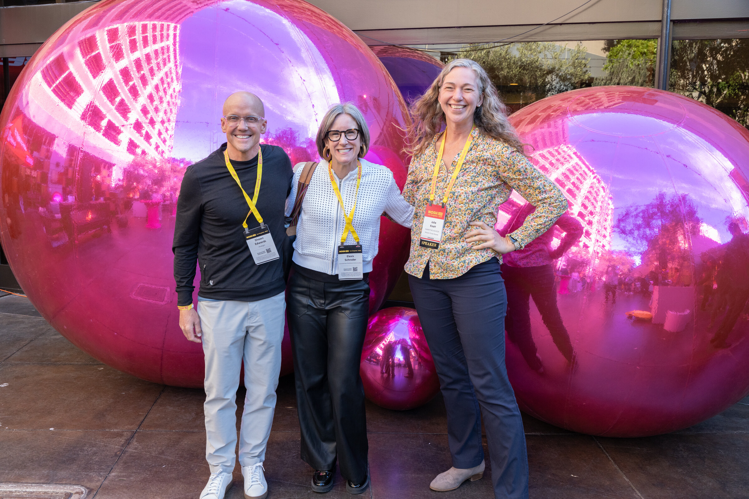 Two women and a man in lanyards standing in front of large pink mirrored spheres.