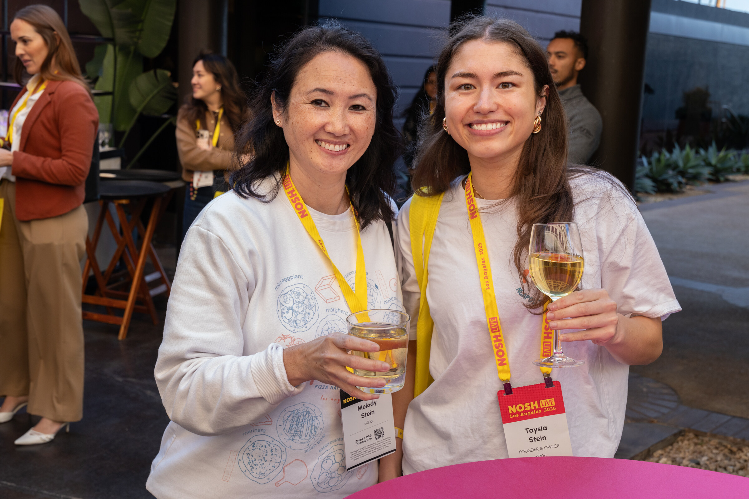 Two smiling women in lanyards holding glasses of white wine at a pink high-top table.