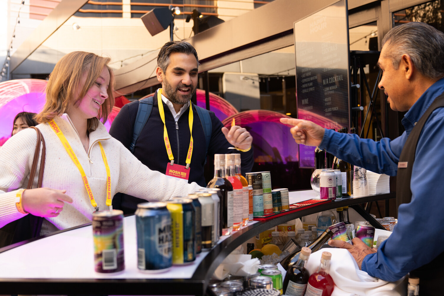 Attendees in lanyards at a bar counter lined with canned beverages while a server points at a menu.