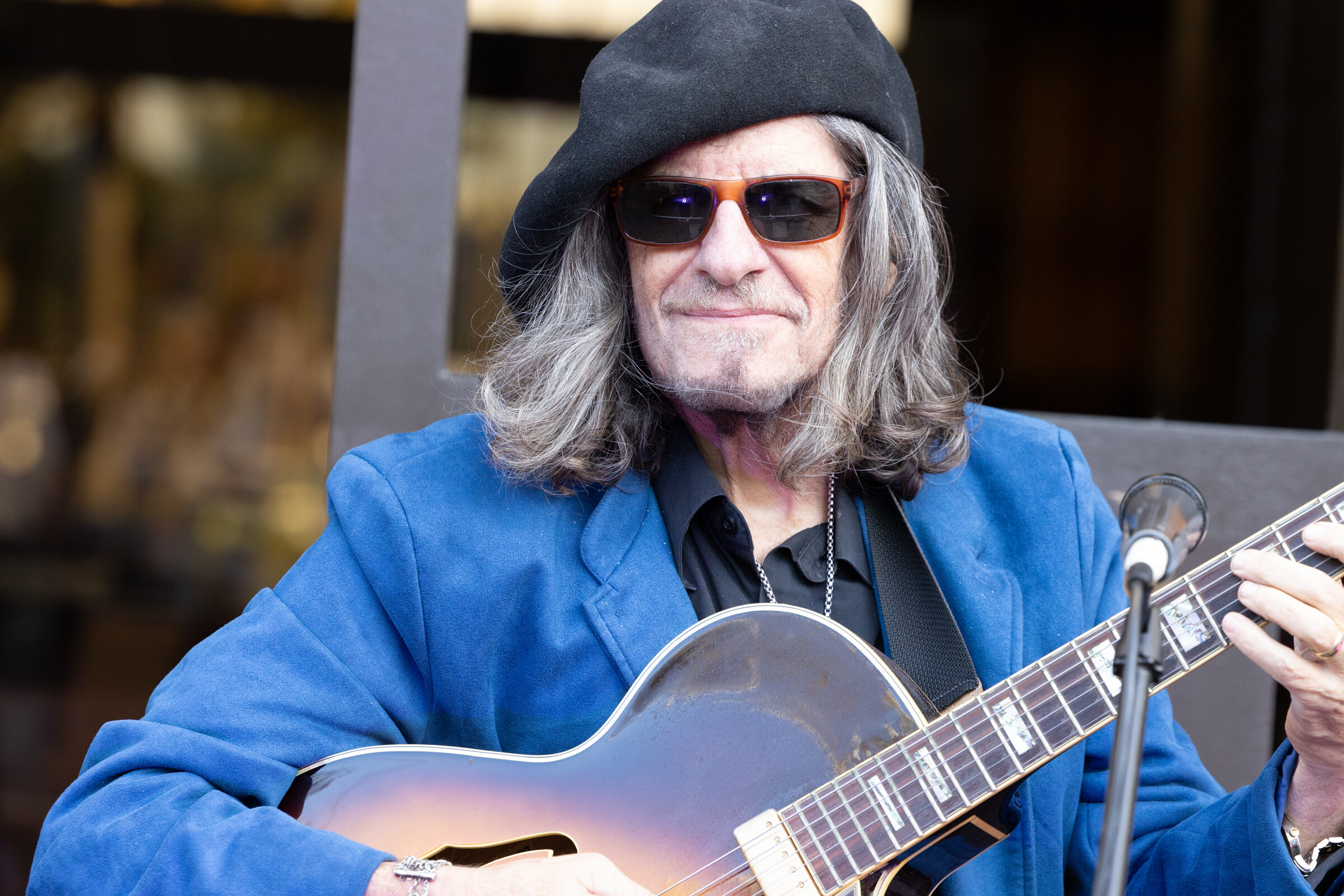 A man in a blue jacket, beret and sunglasses playing an archtop guitar into a microphone.
