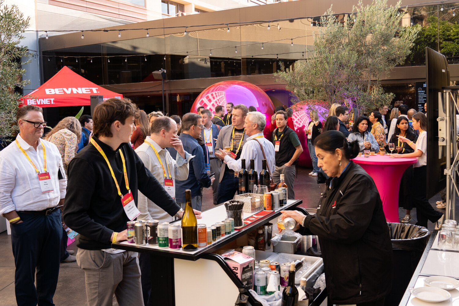 A crowded outdoor courtyard with a bar in front, a red BevNET tent and large pink spheres at the back.