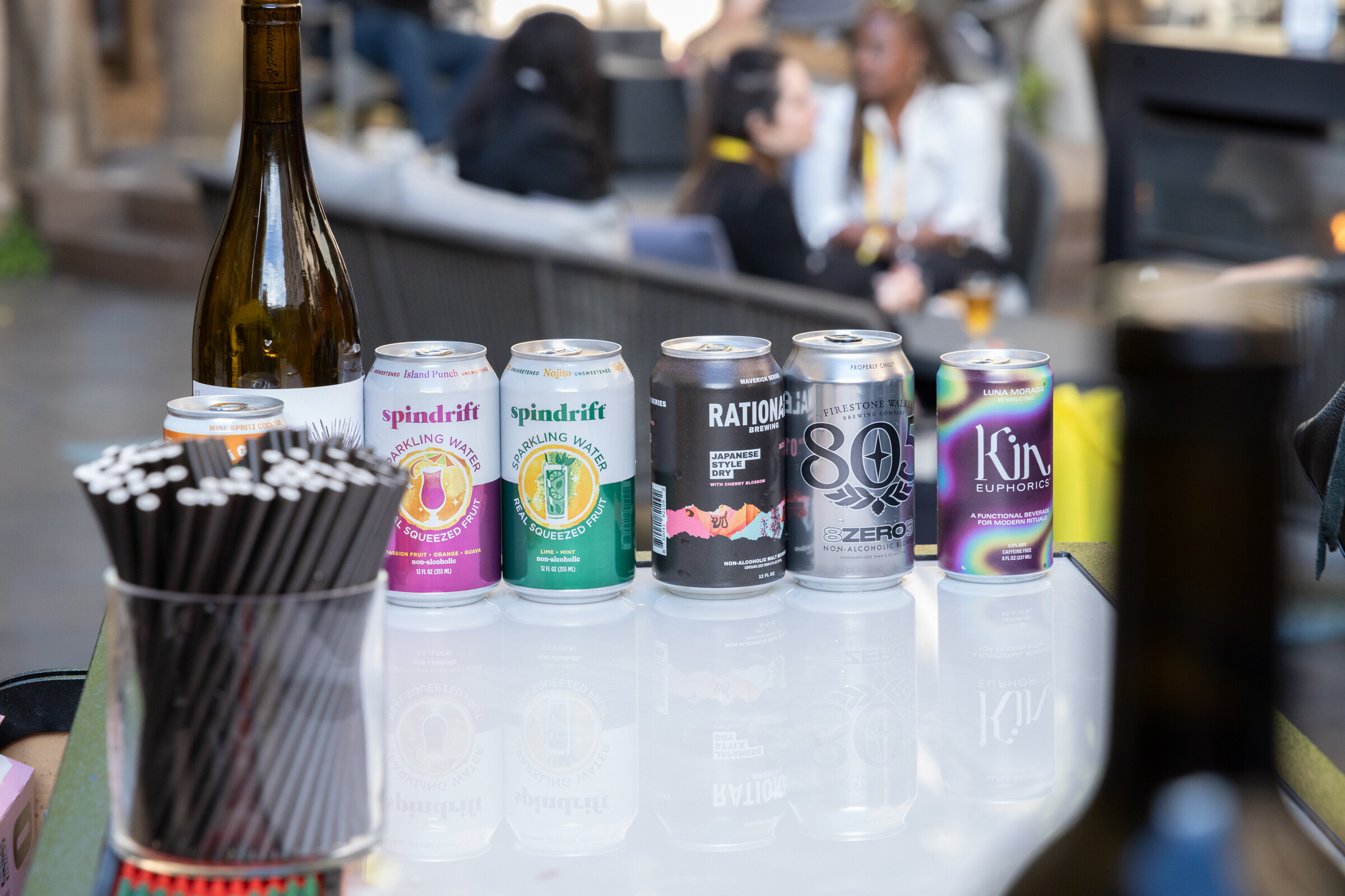 A row of beverage cans including Spindrift, Rationale, 805 and Kin Euphorics next to a wine bottle and a cup of straws.