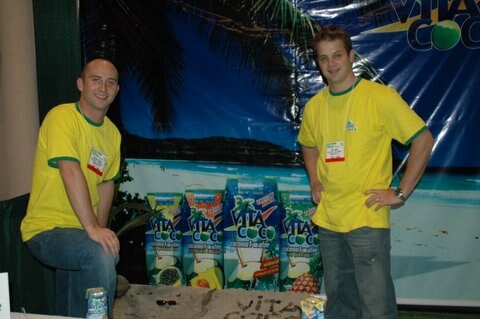Two men in yellow Vita Coco shirts with name badges stand beside cartons of Vita Coco coconut water in front of a beach-scene backdrop with a palm tree.