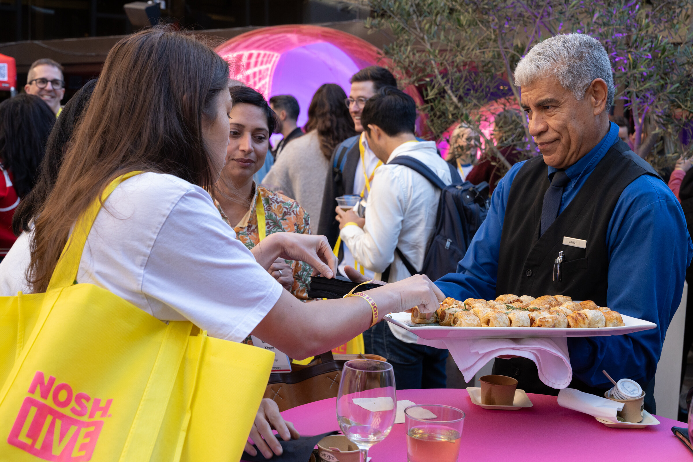 A server in a vest offering a tray of appetizers to attendees beside a pink cocktail table.