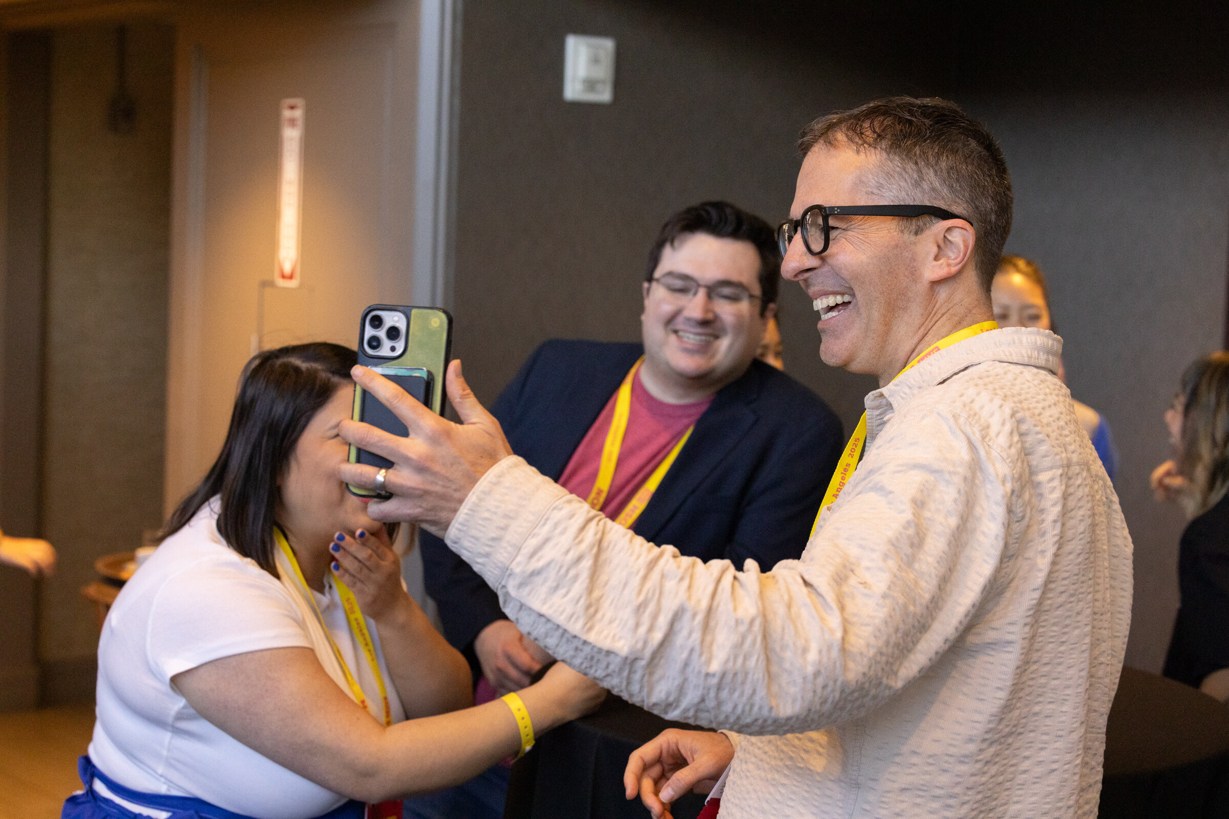 A man in a cream shirt holding up a phone to take a selfie with two others, all with lanyards.