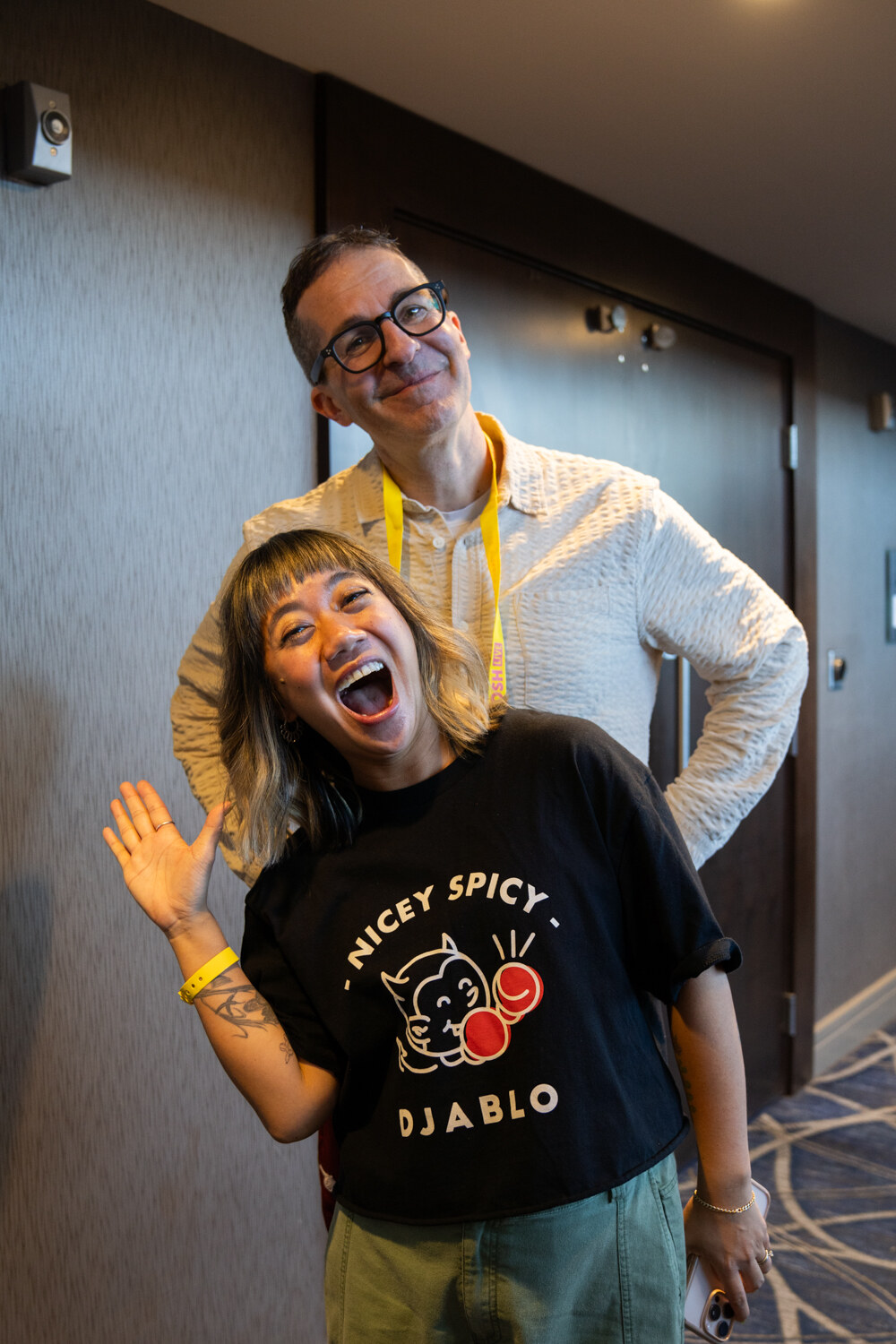 A woman in a black Djablo shirt waving with her mouth open and a man with glasses standing behind her.