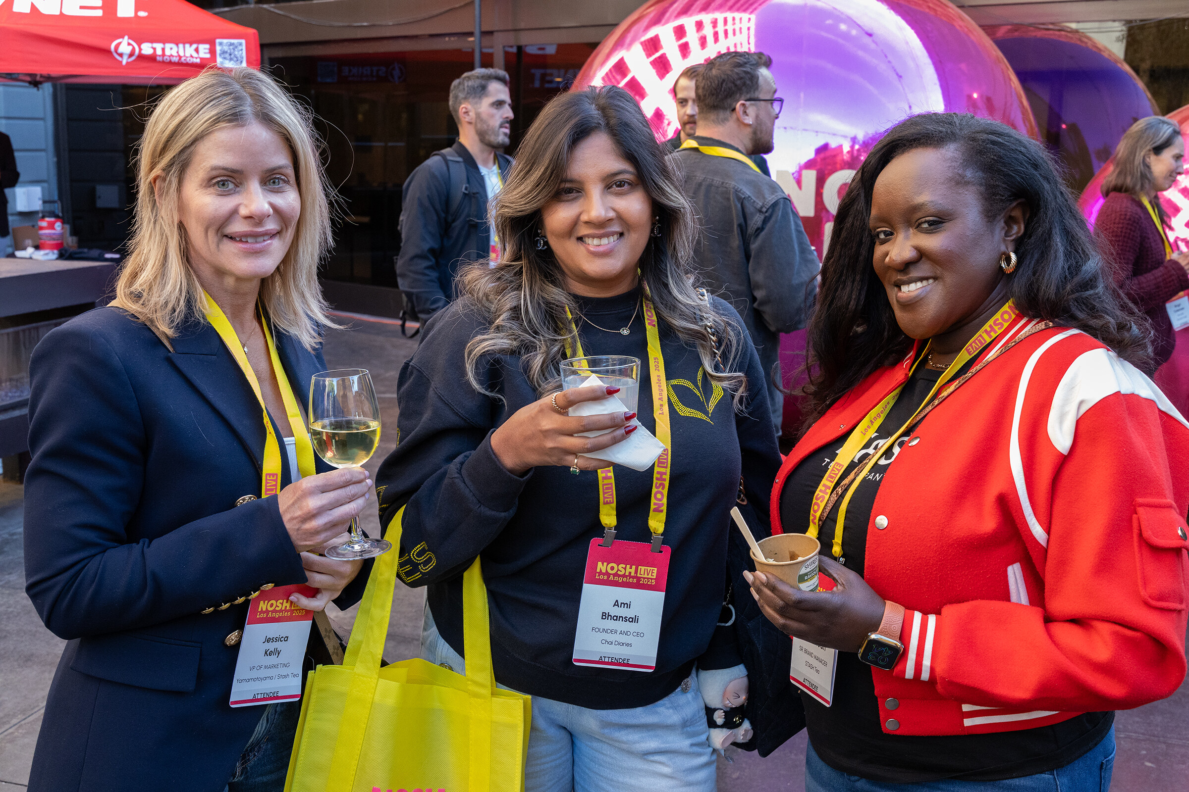 Three smiling women with lanyards, one holding a glass of white wine, standing in the outdoor courtyard.