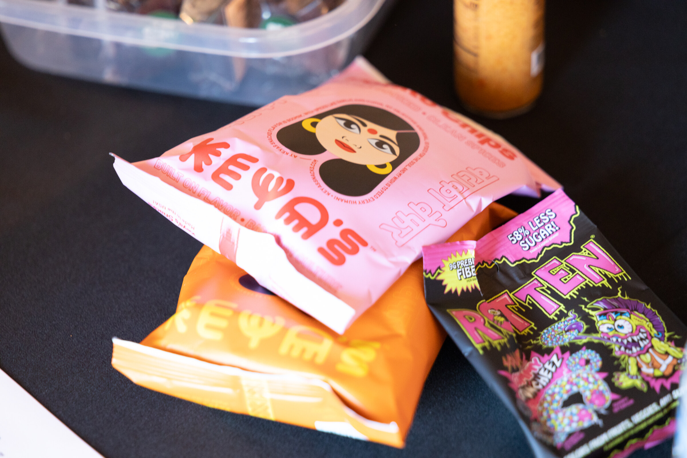 Pink and orange Keya's chip bags next to a black Rotten candy bag on a black tablecloth.