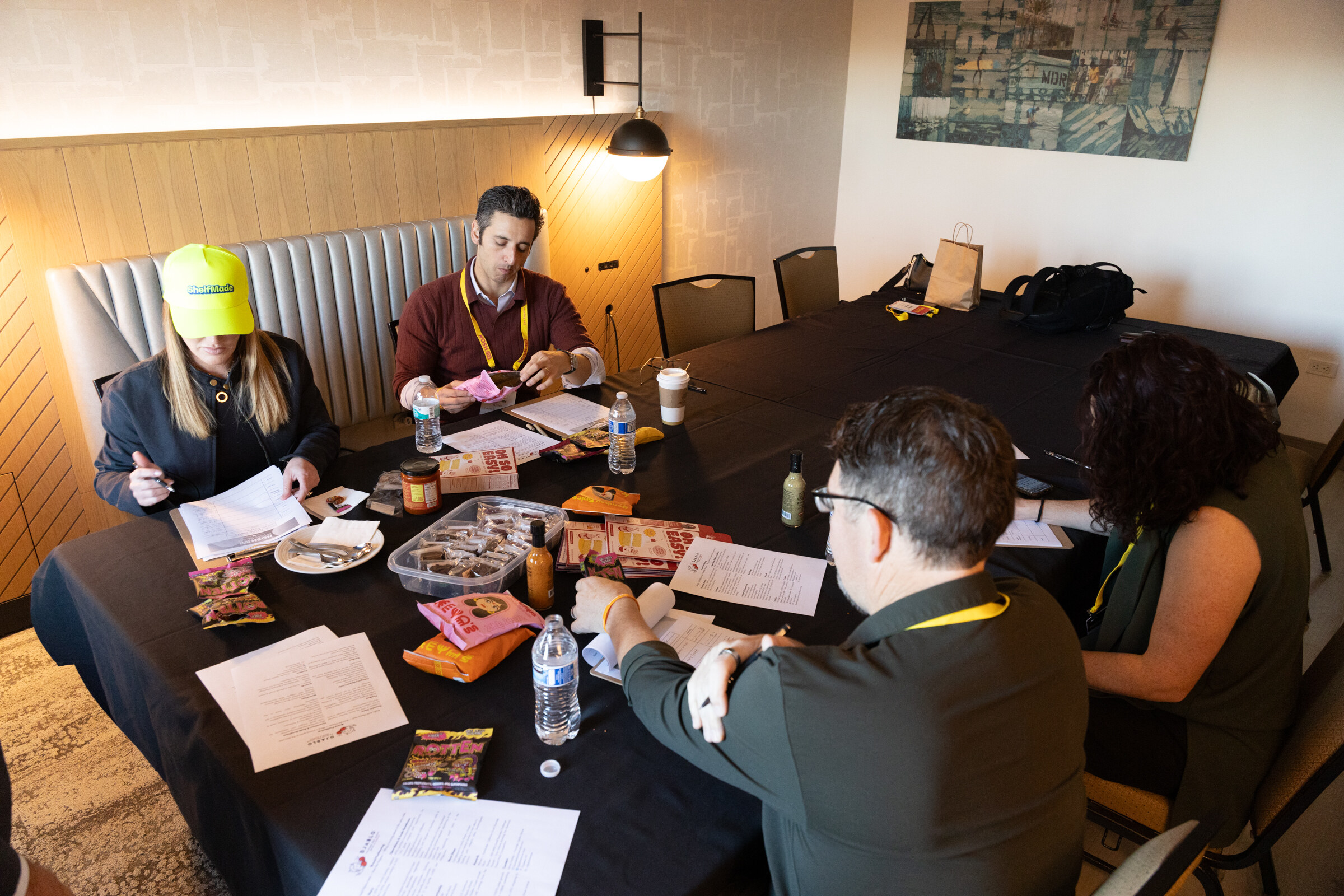Four people seated around a table covered with snacks, sauces and score sheets in a hotel room.