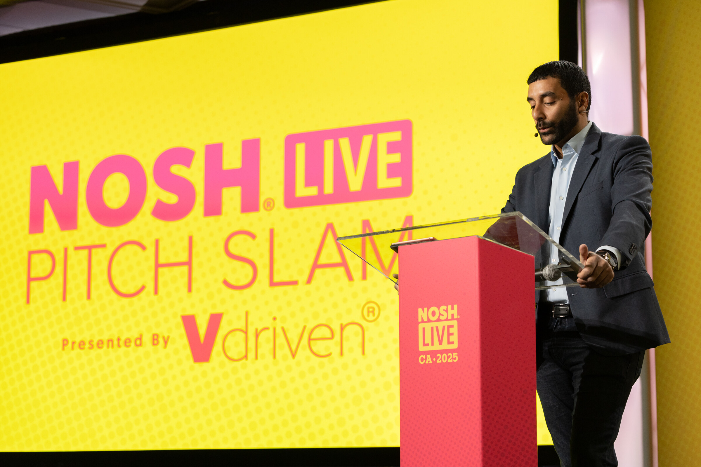 A man in a gray blazer at a clear podium in front of a yellow screen reading Nosh Live Pitch Slam Presented By Vdriven.