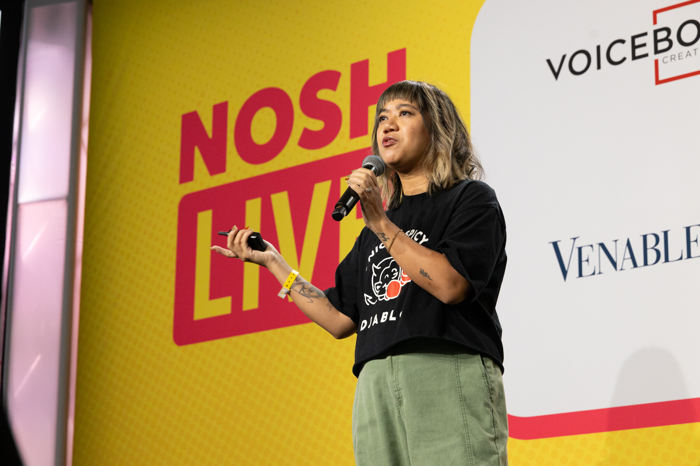 A woman in a black Djablo t-shirt holding a microphone in front of a Nosh Live and Venable backdrop.