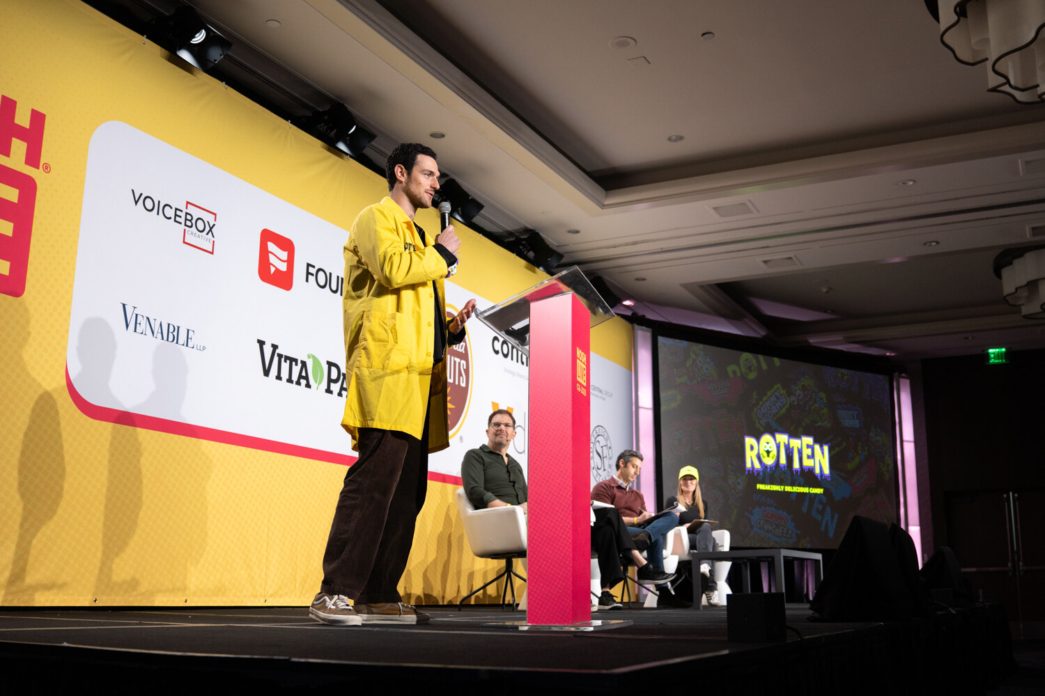 A man in a yellow jacket speaking into a microphone at a podium with a Rotten logo on the screen behind him and judges seated at right.