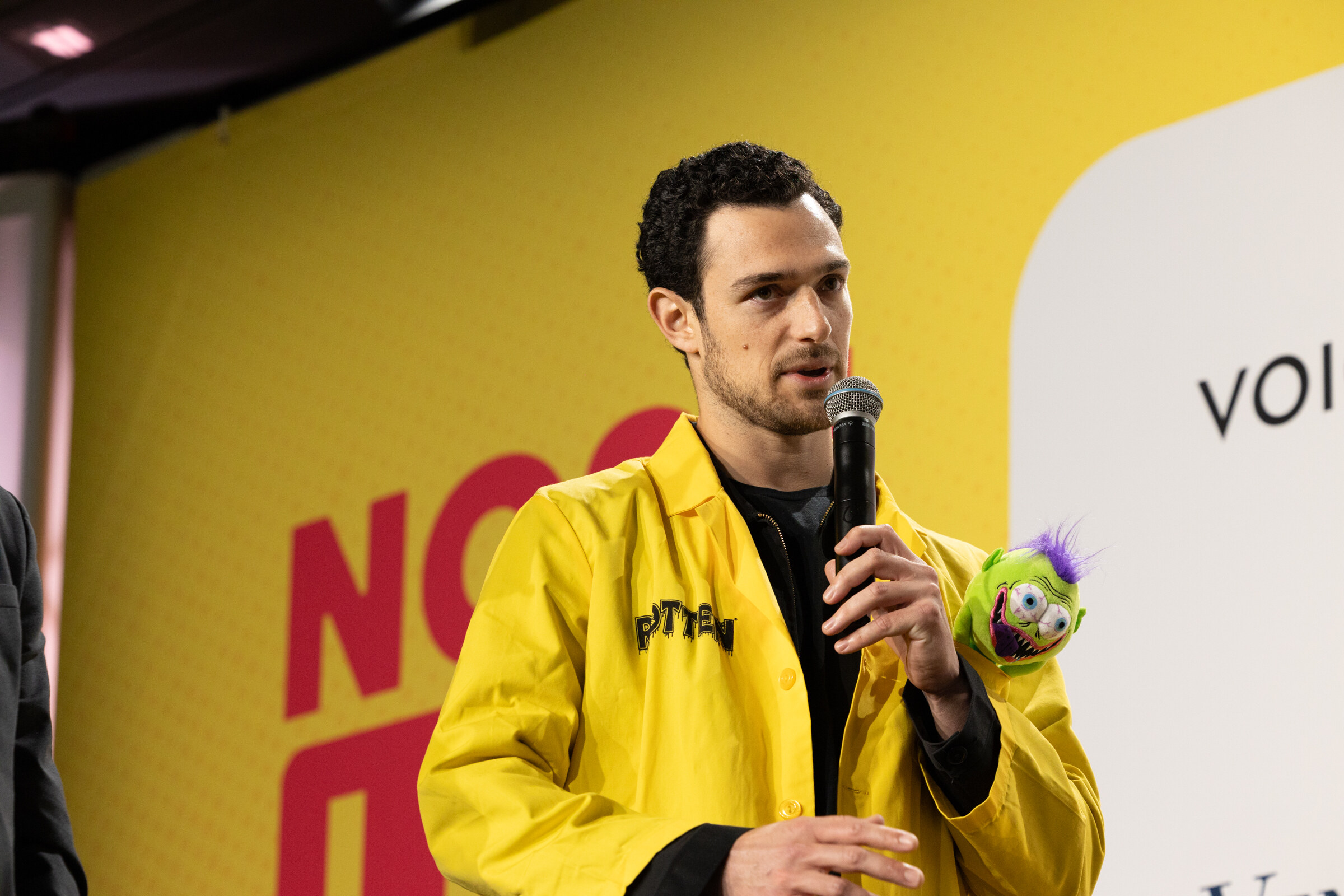 A man in a yellow Rotten jacket speaking into a microphone with a small purple puppet on his shoulder.