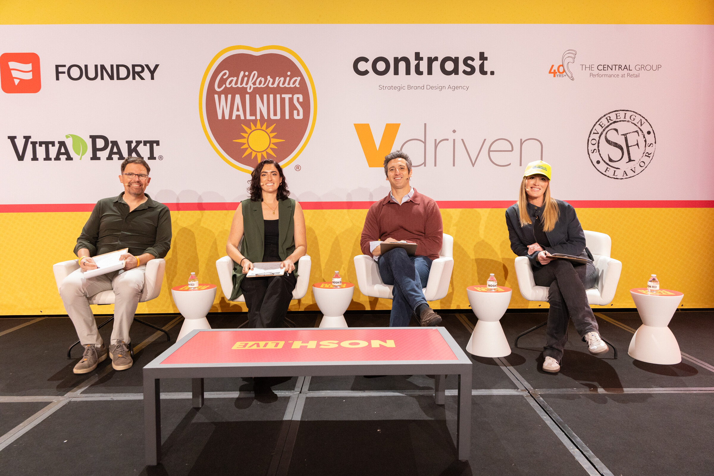 Four judges seated in white chairs on stage in front of a banner with sponsor logos including California Walnuts and Vdriven.