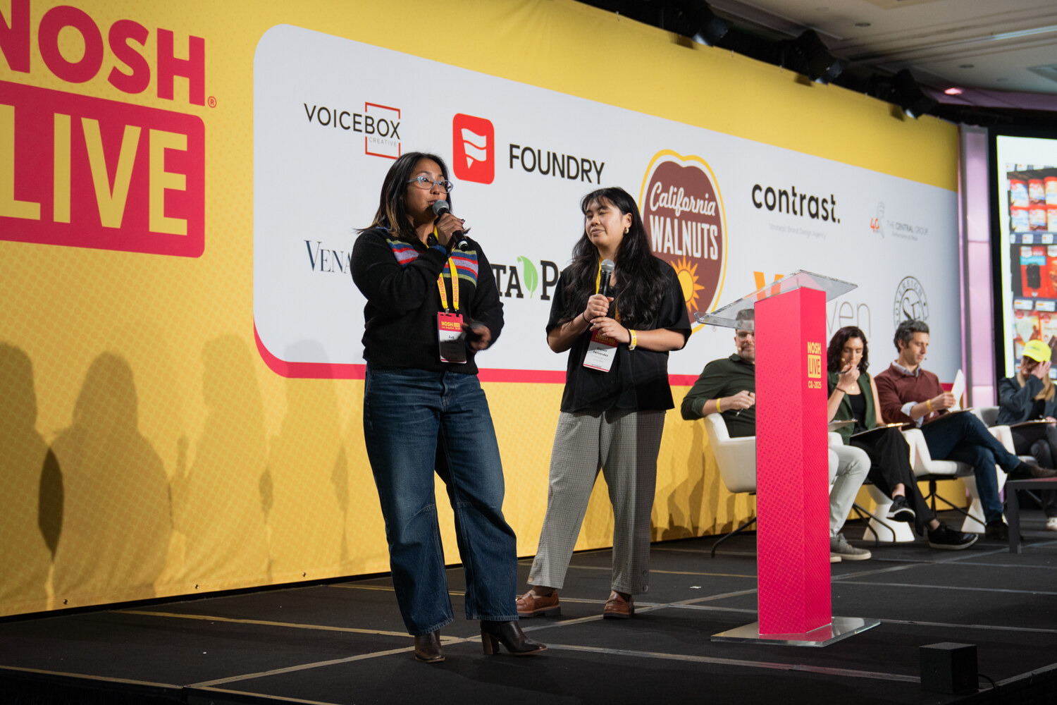 Two women with microphones standing on stage in front of the judges' panel and a Nosh Live backdrop.