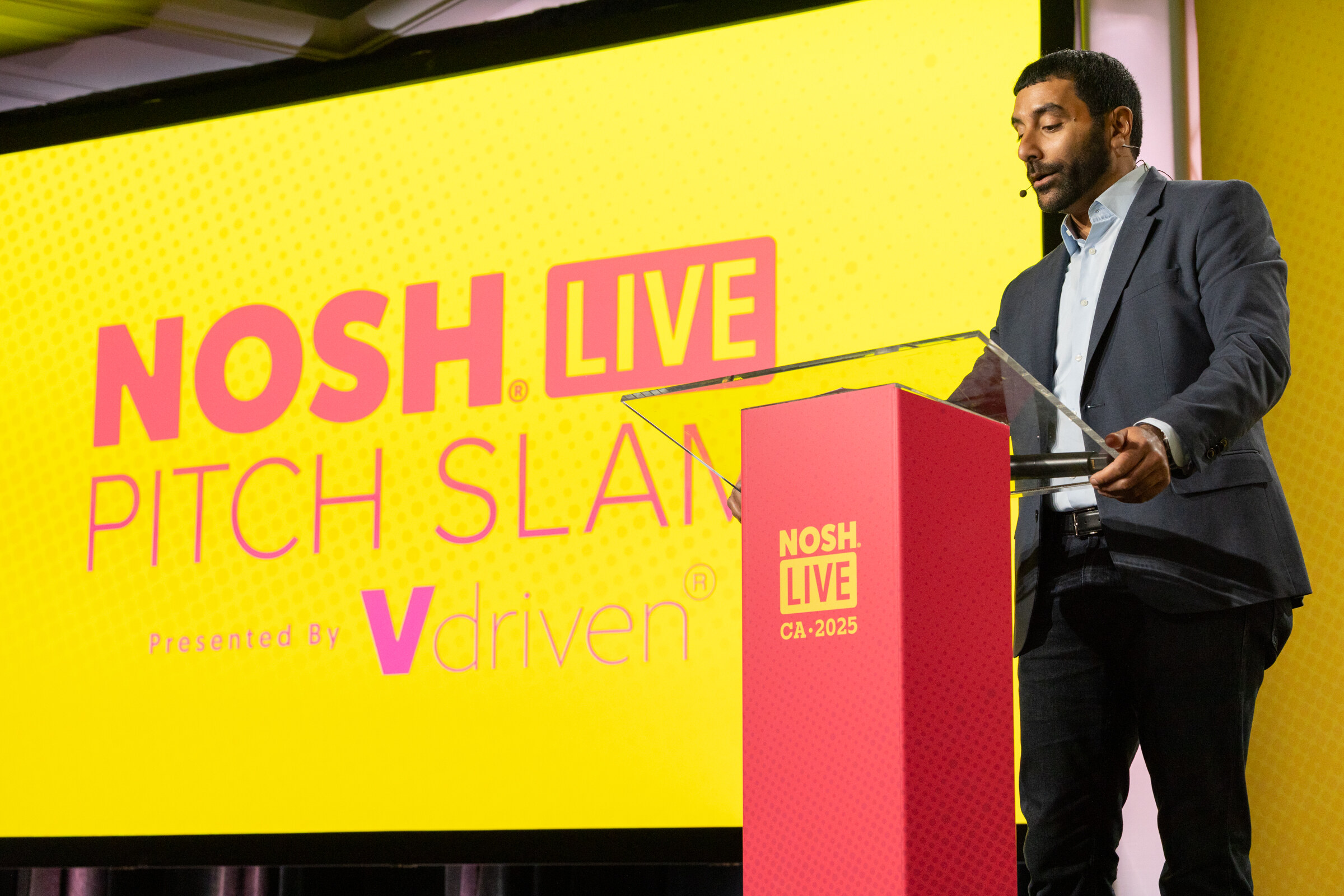 A man in a gray blazer speaking at a podium in front of a yellow Nosh Live Pitch Slam Presented By Vdriven screen.