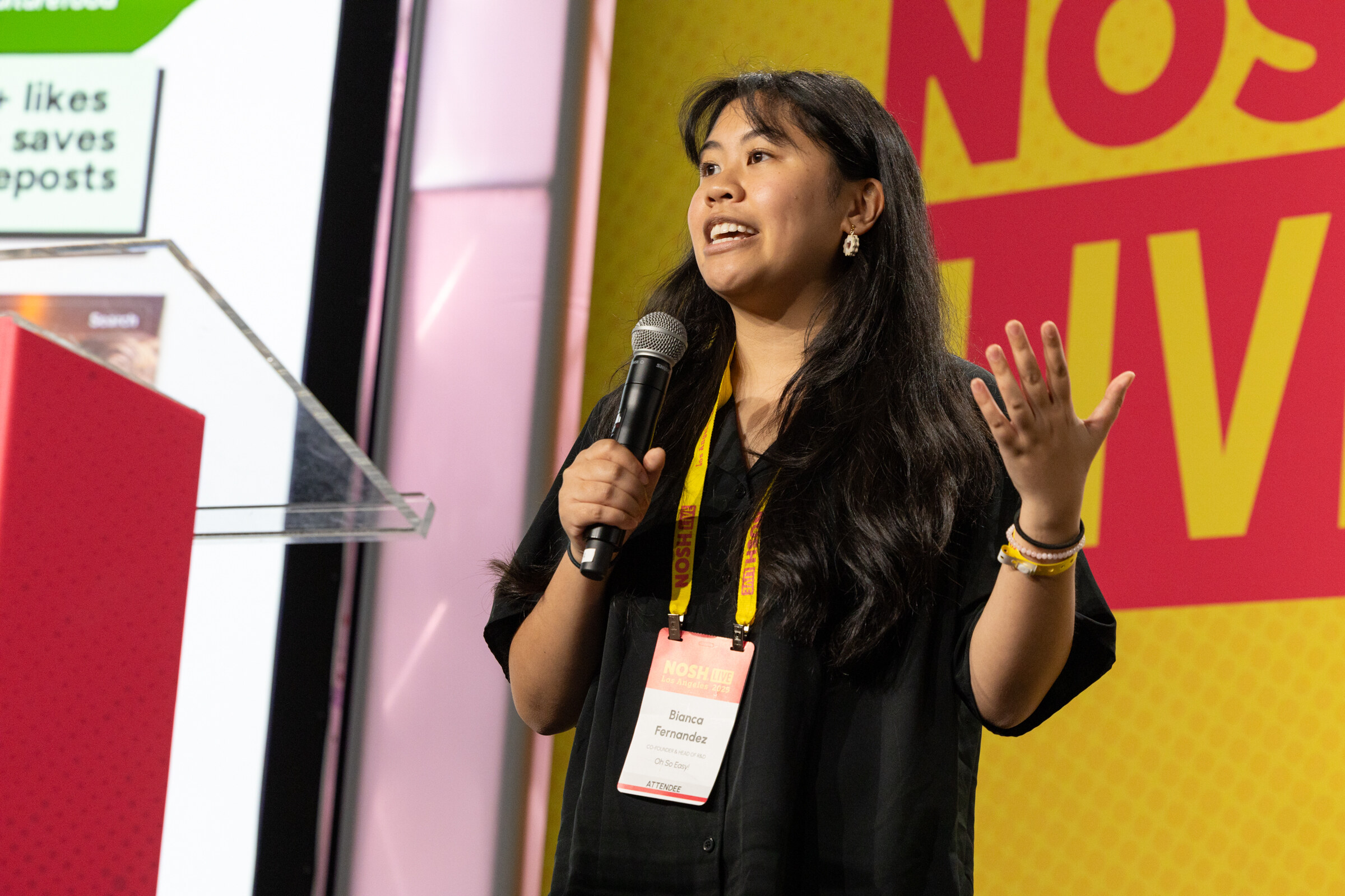 A young woman with long dark hair wearing a badge and speaking into a microphone in front of a Nosh Live backdrop.