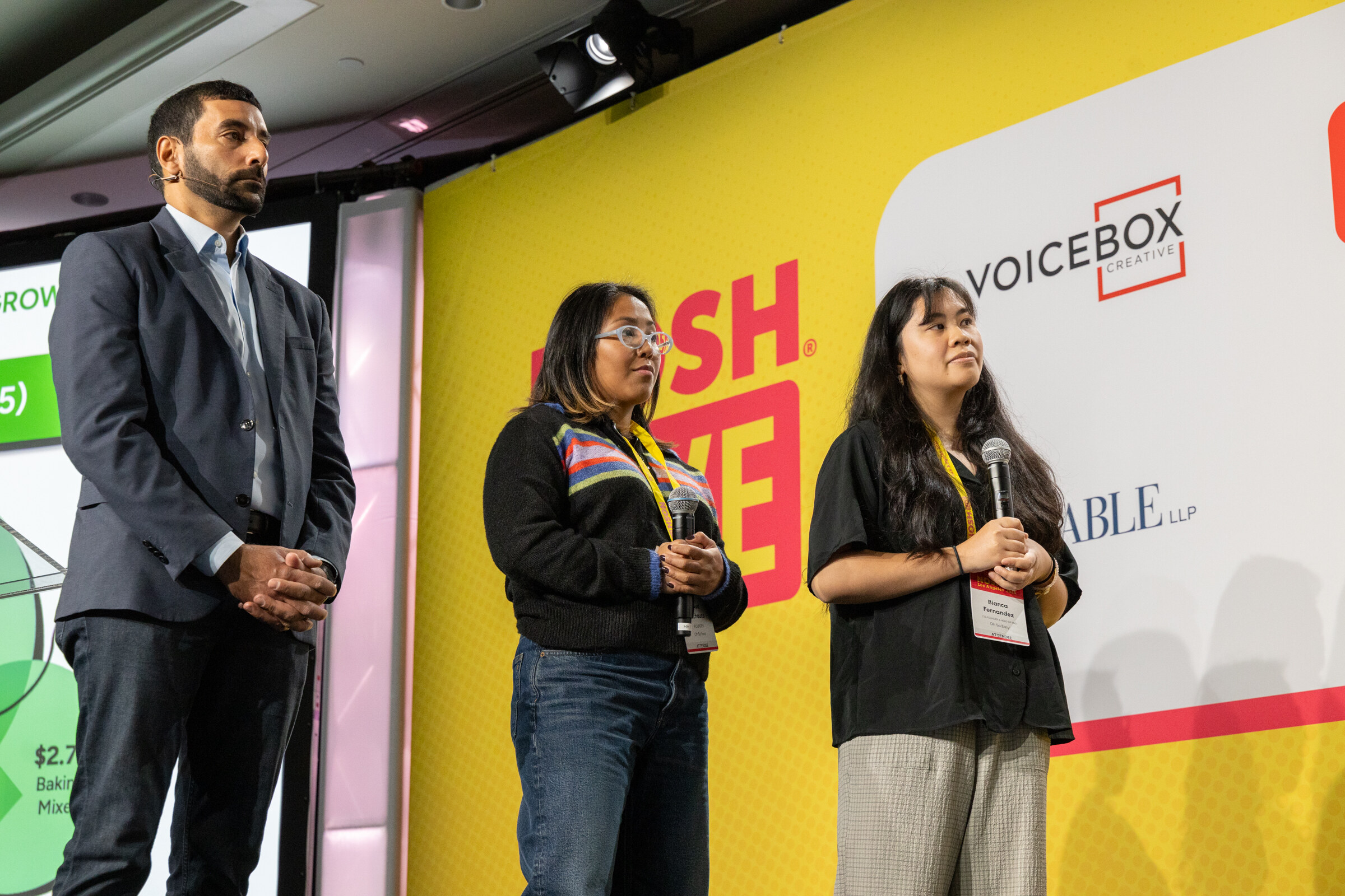 A man in a gray blazer and two women with microphones standing on stage in front of a Nosh Live backdrop.
