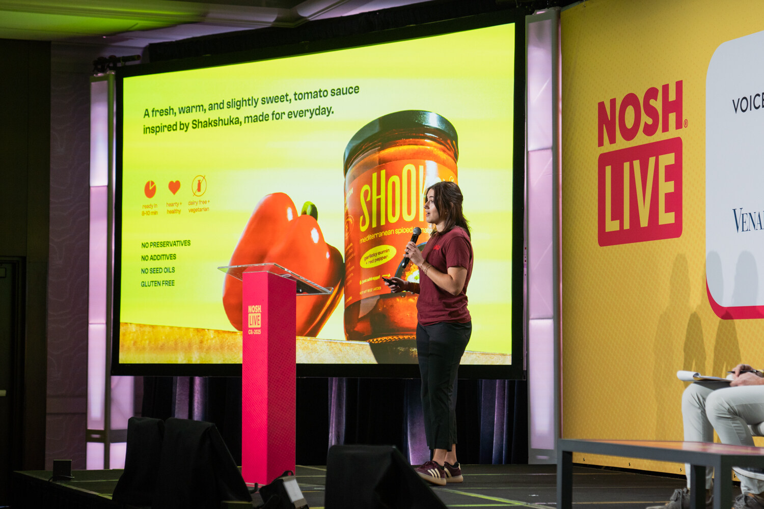A woman with a microphone beside a podium and a large screen showing a jar of Shooka sauce.