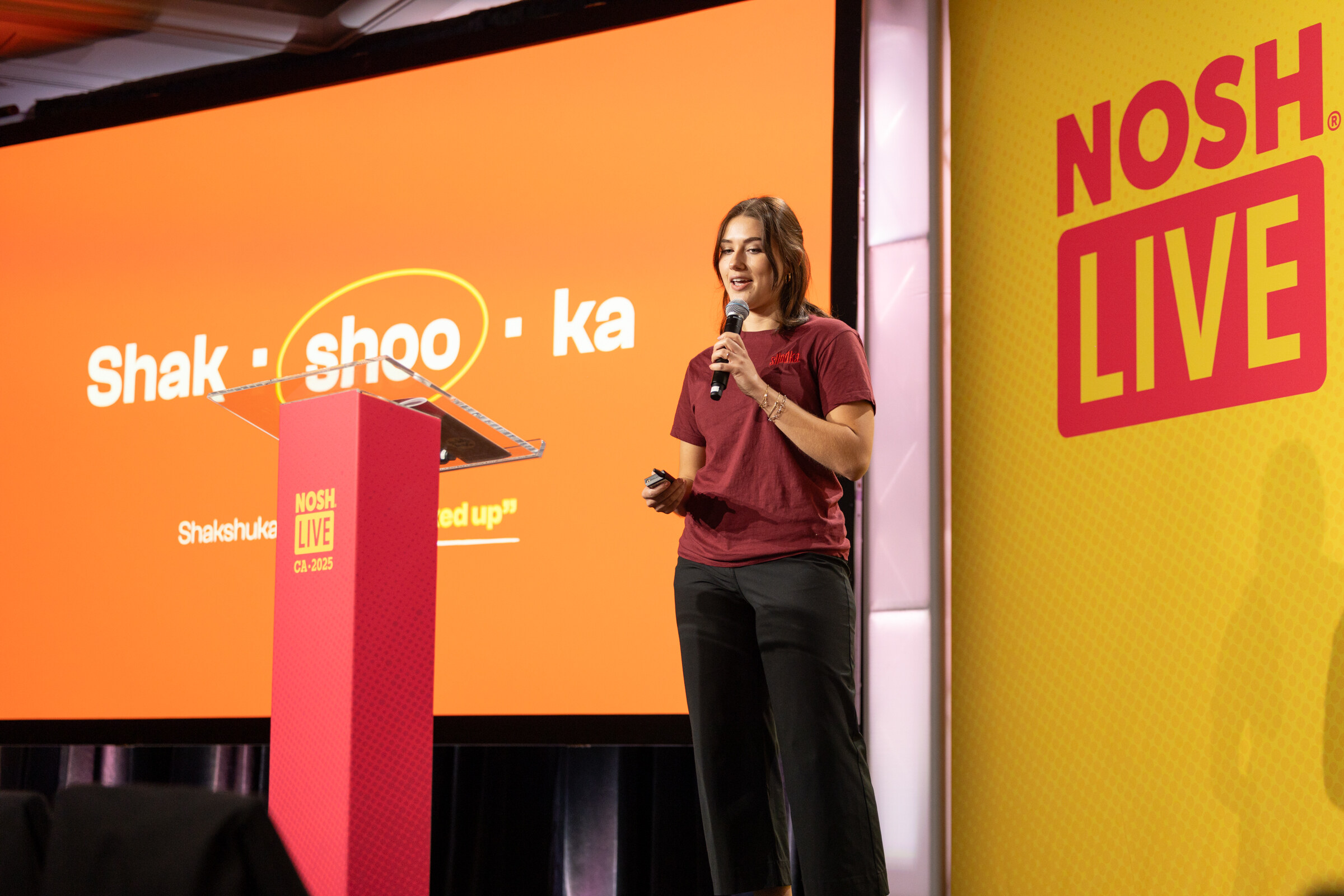 A young woman with a microphone beside a podium in front of an orange slide spelling Shak-shoo-ka.