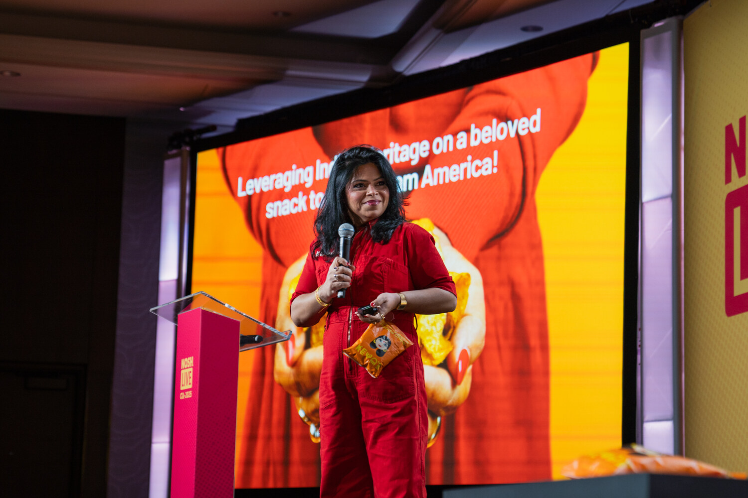 A woman in a red jumpsuit holding a microphone and a snack bag in front of a large orange slide.