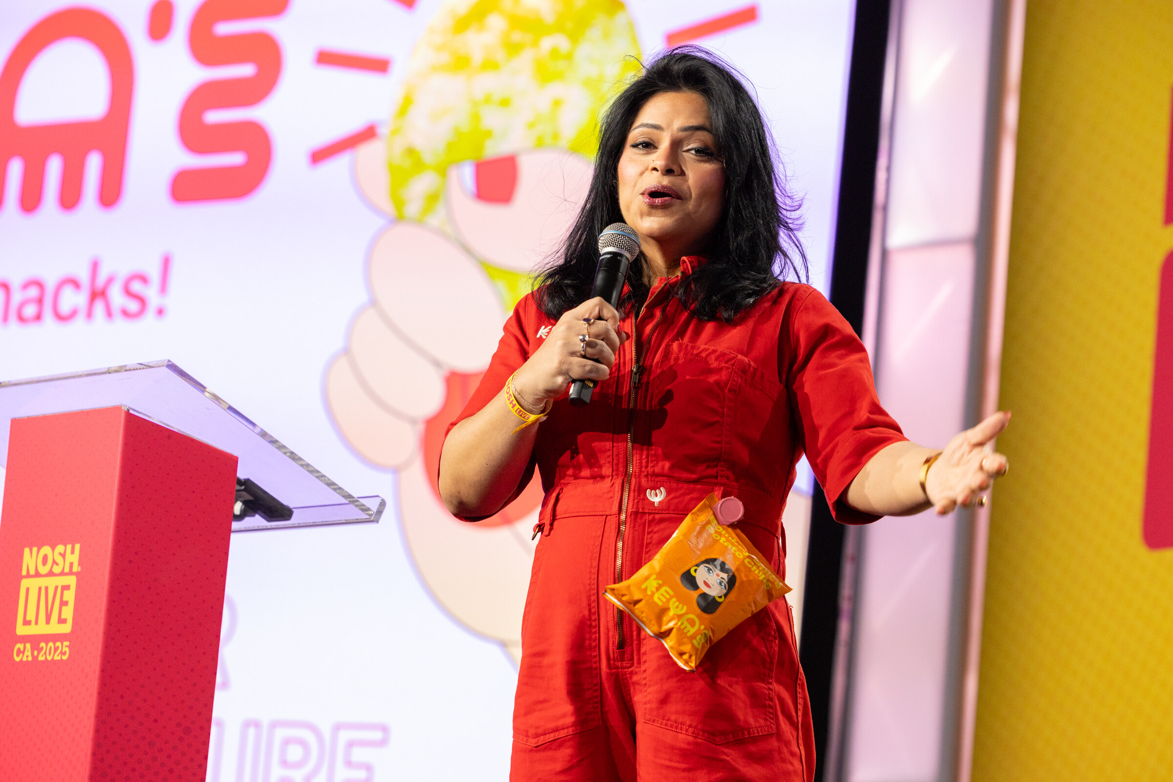 A woman in a red jumpsuit with a microphone and a yellow snack bag in front of a bright snack-themed slide.