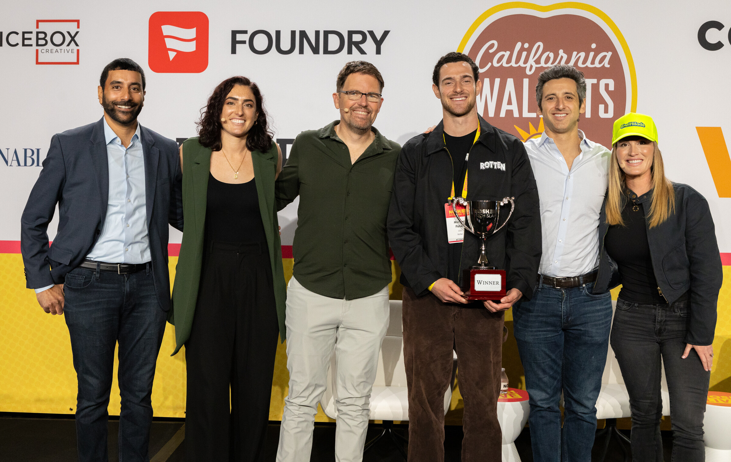 Six people standing in a row on stage, one holding a silver trophy, in front of Foundry and California Walnuts signs.