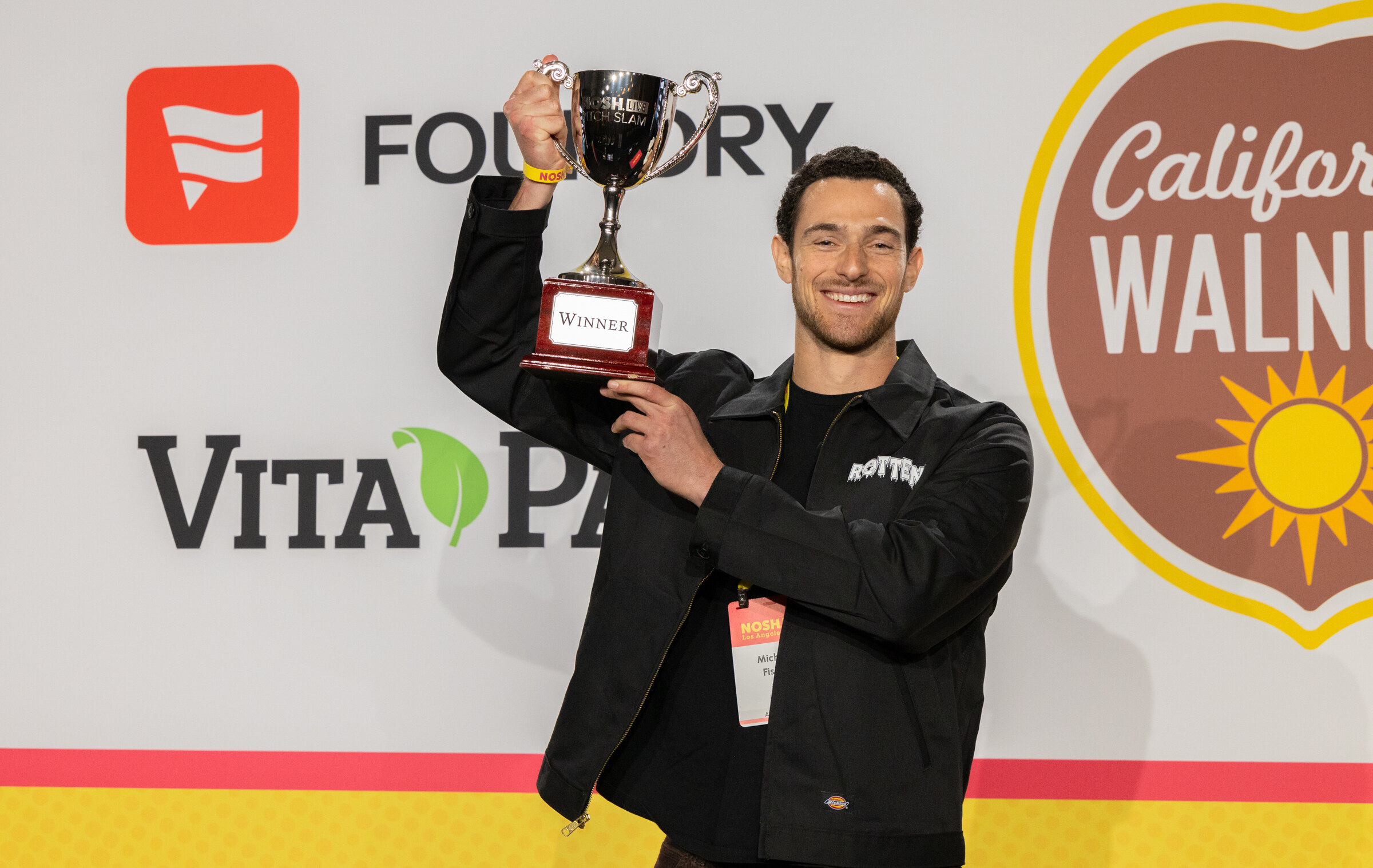 A man in a black jacket marked Rotten smiling and holding a silver winner's trophy above his head.
